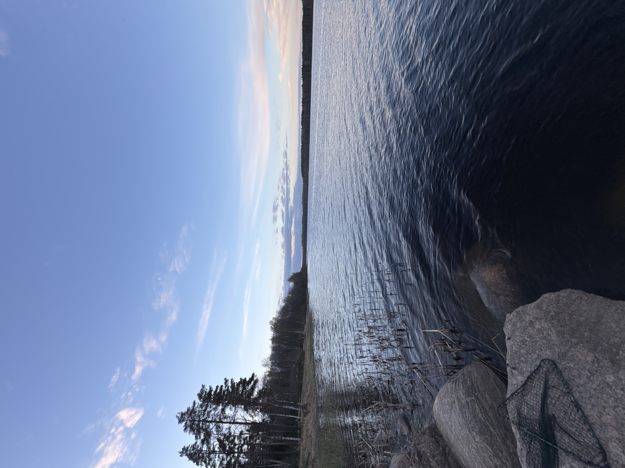 The image depicts a lake scene. The sky is a clear blue with some streaks of white clouds. The lake water is dark blue and slightly choppy. The shoreline is lined with trees and reeds. Large gray rocks are present at the edge of the lake, and a fishing net is draped over one of them. The location is unspecified, and there's no indication of the city Aleksandrovka, Russia. No people are visible.