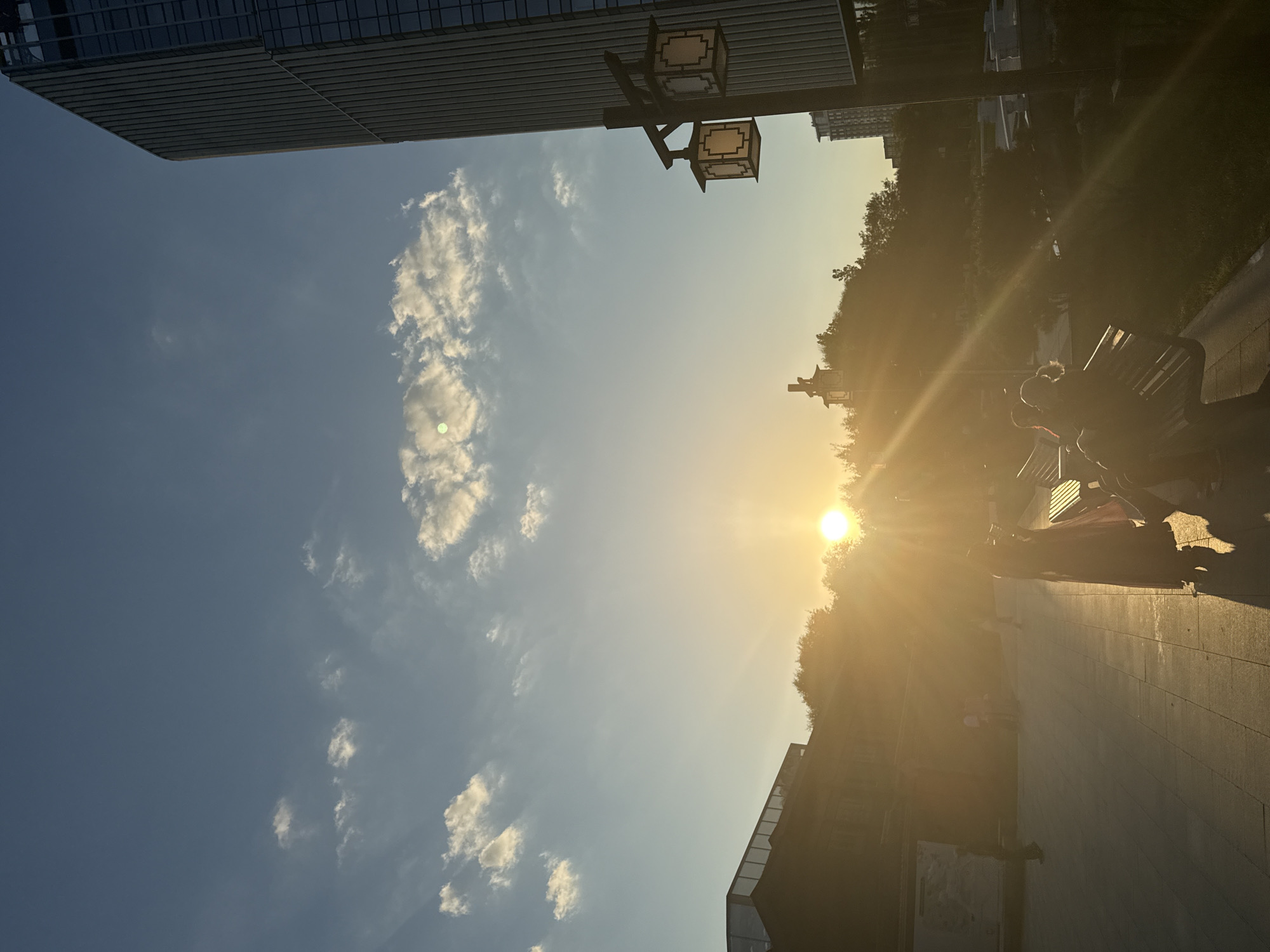 The image depicts a sunset scene in a city, with a modern building on the right and a lower, possibly older, structure to the left. The sun is low on the horizon, casting a warm, golden light and creating lens flare effects. Silhouettes of people are visible, some standing and others sitting on a bench. Two traditional-style lanterns hang from a lamppost. The sky is blue with scattered clouds. The scene appears to be an outdoor public space, possibly a park or plaza, with a paved walkway. The architectural styles suggest a blend of modern and traditional elements within an urban setting. There is no indication that this location is in Moscow, Russia.