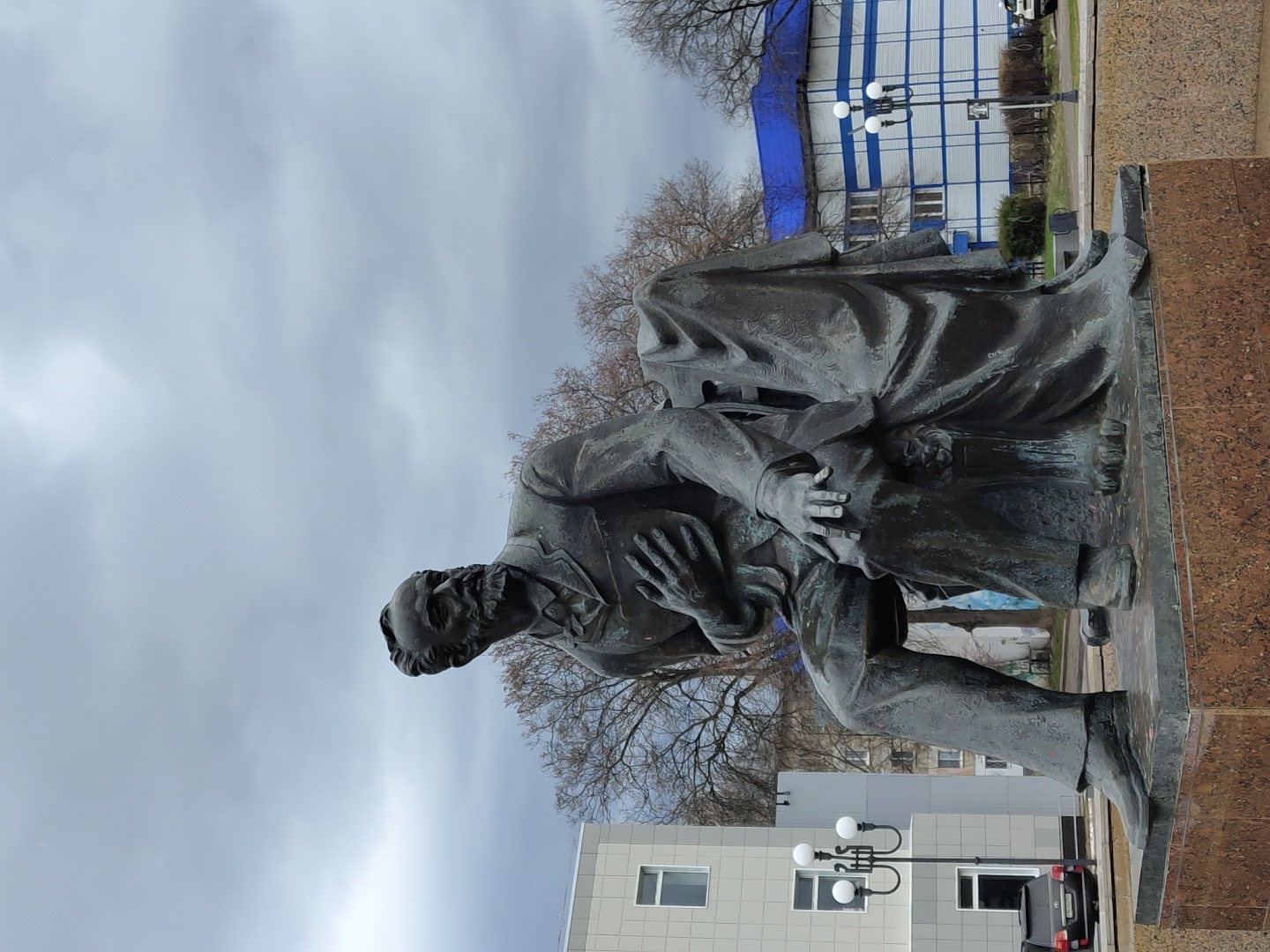A bronze statue of a seated man dominates the foreground of this outdoor urban scene, likely located in Chaikovsky, Russia. The statue depicts a mature man with a prominent beard and mustache, dressed in a formal suit with a long coat, his left hand resting on his chest or stomach, and his right hand on his lap. He appears to be in a contemplative pose. The dark, possibly oxidized, bronze statue rests on a polished reddish-brown granite plinth.
The scene is set under an overcast, cloudy sky, suggesting a cool day, possibly in autumn or winter given the bare branches of the deciduous trees visible in the midground and background. In the background, there are various buildings, including one with distinctive horizontal white and blue siding, and another modern grey building with visible windows. A black lamppost with multiple white globe lights stands near the blue and white building. A dark-colored car is partially visible, parked near the grey building in the lower right corner. The overall setting is a public space, such as a square or park, typical for monuments in a city. No visible text is discernible on the statue or its plinth, nor on any surrounding objects or buildings.
