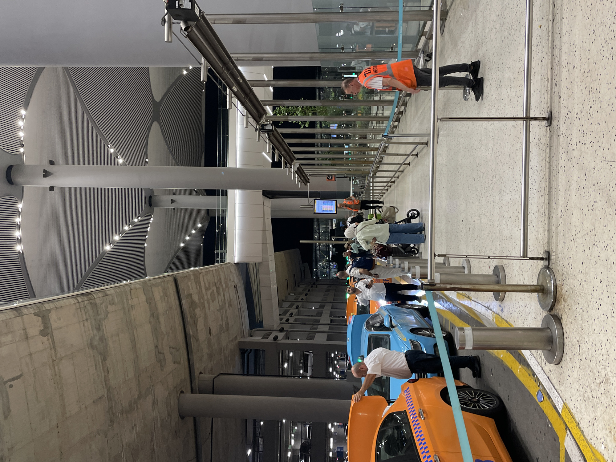 An exterior shot shows a queue of taxis and people at what appears to be an airport in Arnavutköy, Turkey. The yellow and blue taxis are lined up, with their trunks open and drivers interacting with passengers or staff. People are standing in line, some with luggage, and a man in an orange vest is directing the flow of traffic. The architecture of the building is modern, with a large, curved ceiling and exposed concrete elements. The lighting suggests it is either evening or night.