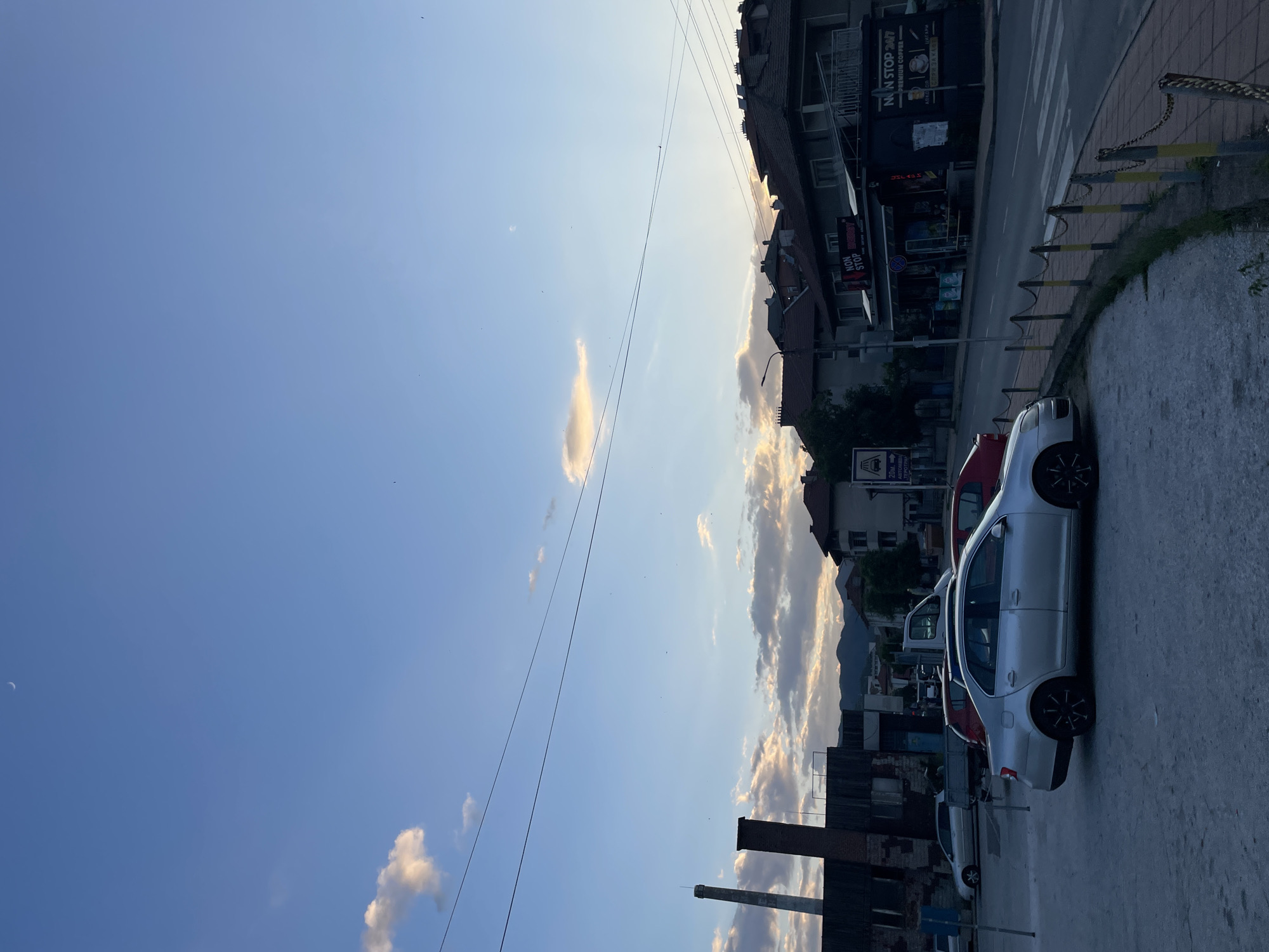 The image shows a street scene in Zlatograd, Bulgaria. The sky is mostly clear blue with a few scattered clouds. Buildings line the street, including a building with a "NON STOP 24/7" sign. There are several parked cars, including a silver sedan in the foreground. In the distance, there are mountains. Power lines cross the sky. A partially ruined brick structure is in the lower left.