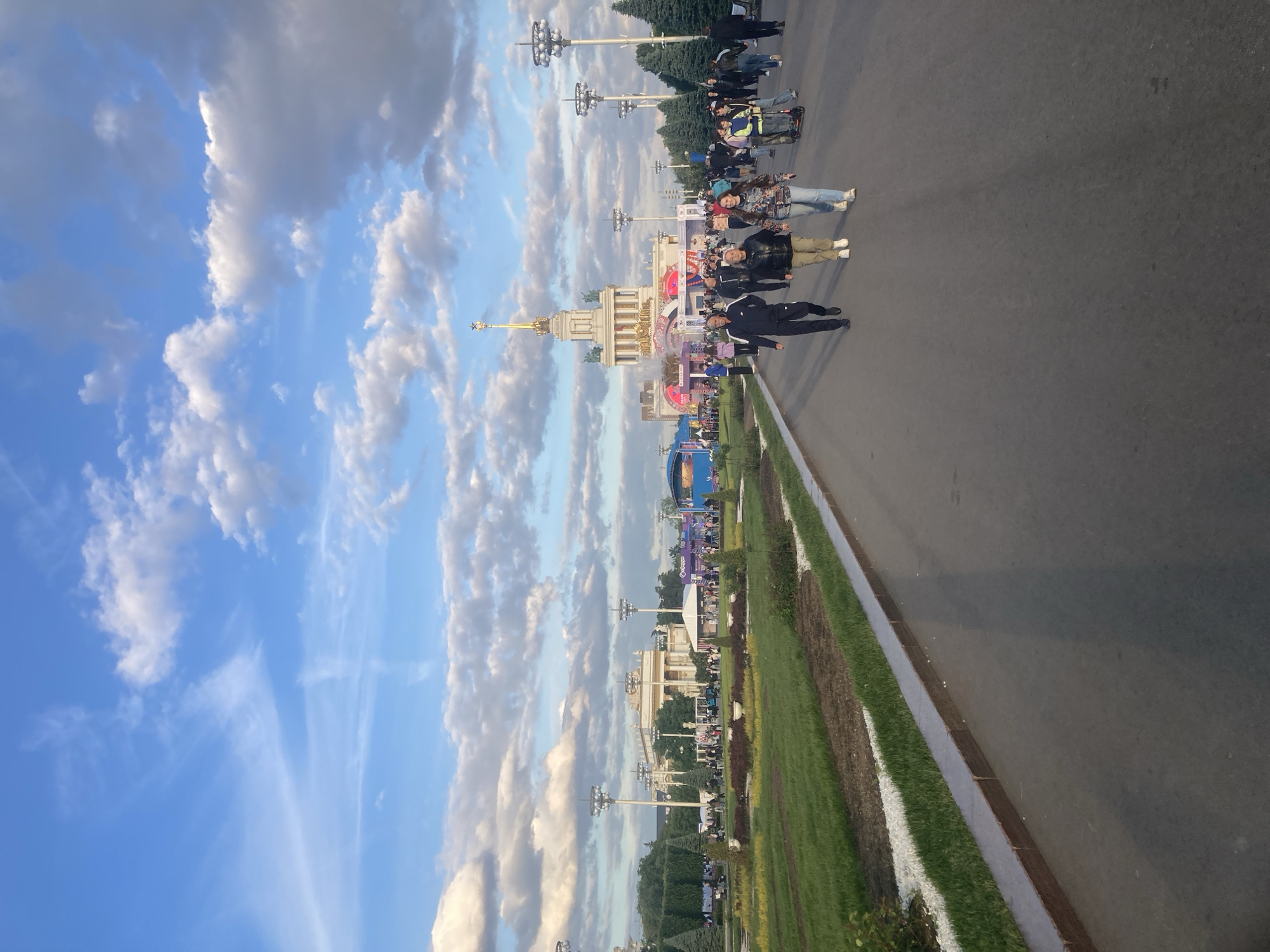 The image captures an outdoor scene at what appears to be a large exhibition or park complex, characteristic of VDNKh in Moscow, Russia. The perspective is from a paved path or road, looking towards a grand architectural structure and a gathering of people.
In the mid-ground, a prominent ornate building with a light-colored facade, intricate detailing, and a golden spire stands out. Flags and banners, some red with white text that is difficult to decipher, adorn its front. Surrounding this building and stretching into the background are other structures, some with colorful signs, and numerous tall, multi-headed lampposts. Beyond these, a line of dark green trees forms a backdrop.
Several people are visible on the paved area. In the foreground, a person in a dark jacket and trousers walks away from the camera. To their left, another individual in a black jacket and tan pants stands facing the camera. Further back, numerous people are gathered around the main building and other structures, suggesting a casual visit or event.
To the left of the paved area, a long, well-maintained strip of grass and some flowerbeds or low planting is visible, bordered by a concrete curb. The sky above is bright blue with a mix of fluffy white cumulus clouds and thinner cirrus clouds, indicating daytime, likely late afternoon or early evening given the soft quality of light and the shadows. The overall atmosphere appears calm and pleasant. Due to distance and resolution, specific text on banners or signs is largely illegible.