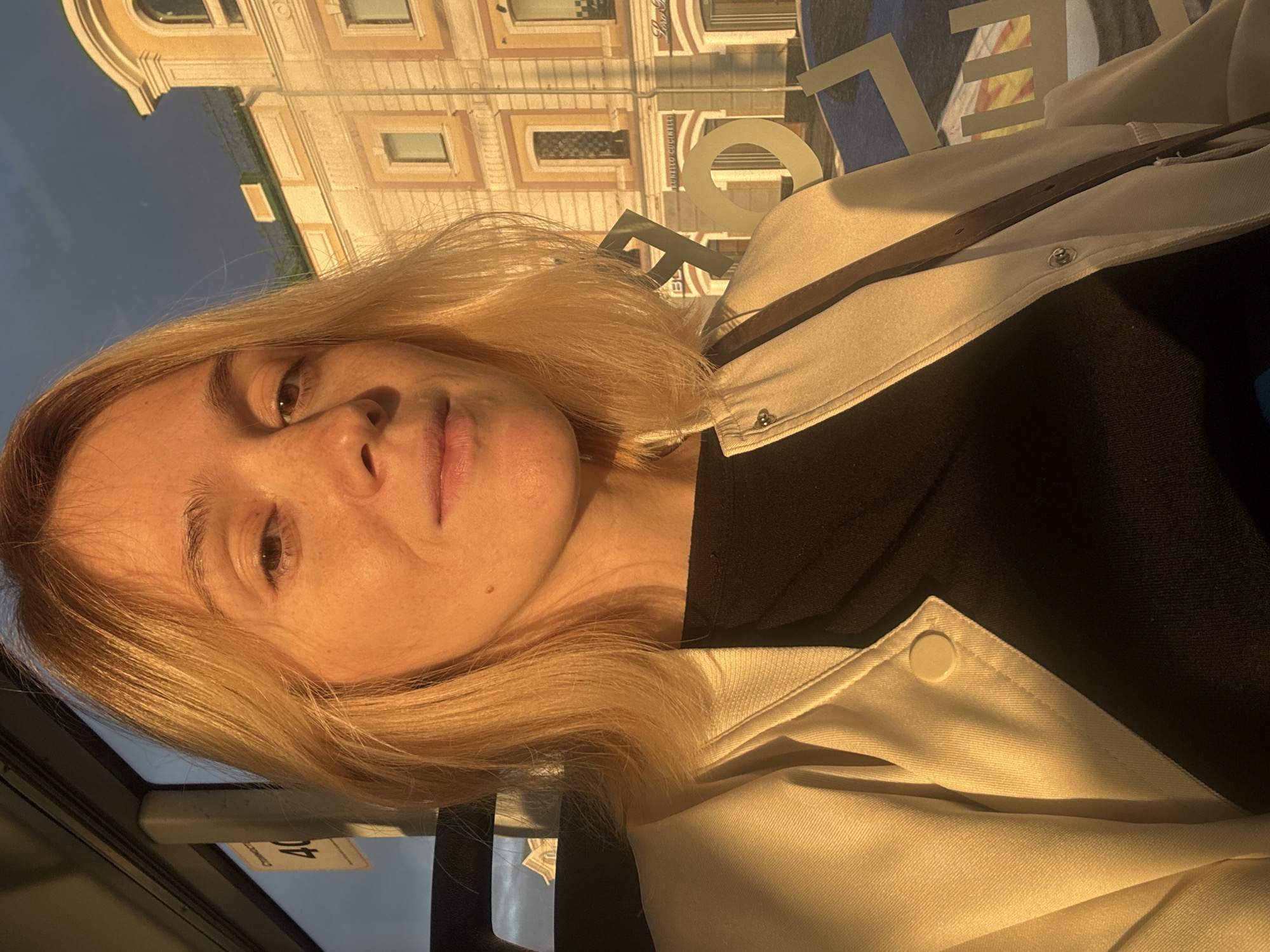 A woman with blonde hair and fair skin is taking a selfie. She is wearing a cream-colored jacket over a black shirt. The sun is setting, casting a warm glow on her face and illuminating the scene. In the background, there is a building with yellow facade and arched windows, which appears to be a historic structure. There is also a sign in the background with the number "40" on it, indicating a route number. The environment suggests the woman is in a public transport vehicle, likely a bus or tram, as a window is visible in the foreground. The reflection on the window shows an urban setting with buildings and signage. Based on the signage and architectural style, the location is likely Nizhny Novgorod, Russia.