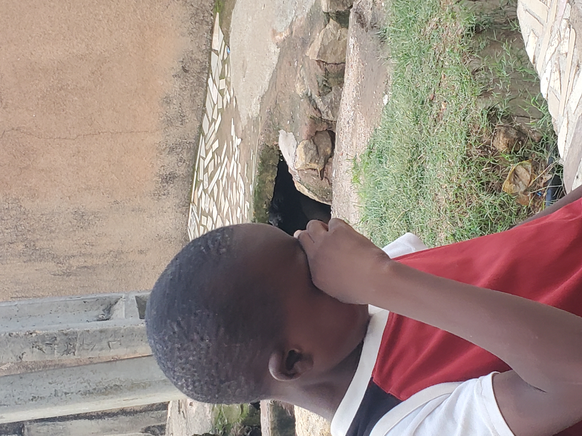 The media file shows a close-up view of a young boy's head and upper body, seen from the side. He is wearing a red and white shirt. His right hand is raised to his face, partially covering his mouth and nose. His eyes are not visible. The boy's skin tone is dark.
The background is partially blurred, but appears to be an outdoor environment. To the left, there is a textured wall with a mosaic-like pattern of small, light-colored rectangular tiles. Further left, there are concrete steps or ledges. In the center and to the right, there is green grass and some dirt with rocks. A dark opening, possibly a drain or small tunnel, is visible amidst the rocks in the center of the image. The lighting suggests it is daytime, with no specific cues for time of day or weather. The mosaic pattern on the wall and the presence of grass and dirt suggest an urban or semi-urban setting, possibly a backyard or a public space in Jos, Nigeria. There is no visible text in the image.