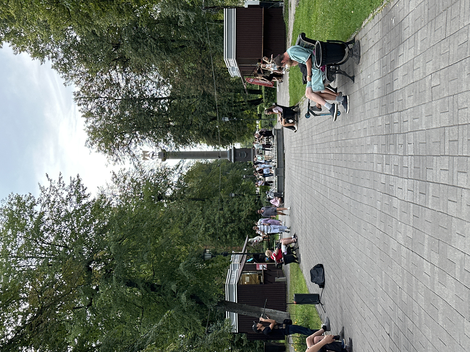 The image depicts a public park with a paved walkway lined by trees on either side. In the foreground, a person is seated on a bench, and another person is standing near a microphone and speaker setup. Further down the walkway, a fountain is visible with people gathered around it. Two small kiosks or stalls are positioned on the left and right sides of the walkway. A tall monument or column with a sculpture on top stands in the distance at the end of the walkway. The scene appears to be set in a city park, possibly in Rusanovo, Russia, given the context provided. The overall atmosphere is relaxed and casual, with people enjoying the park space.