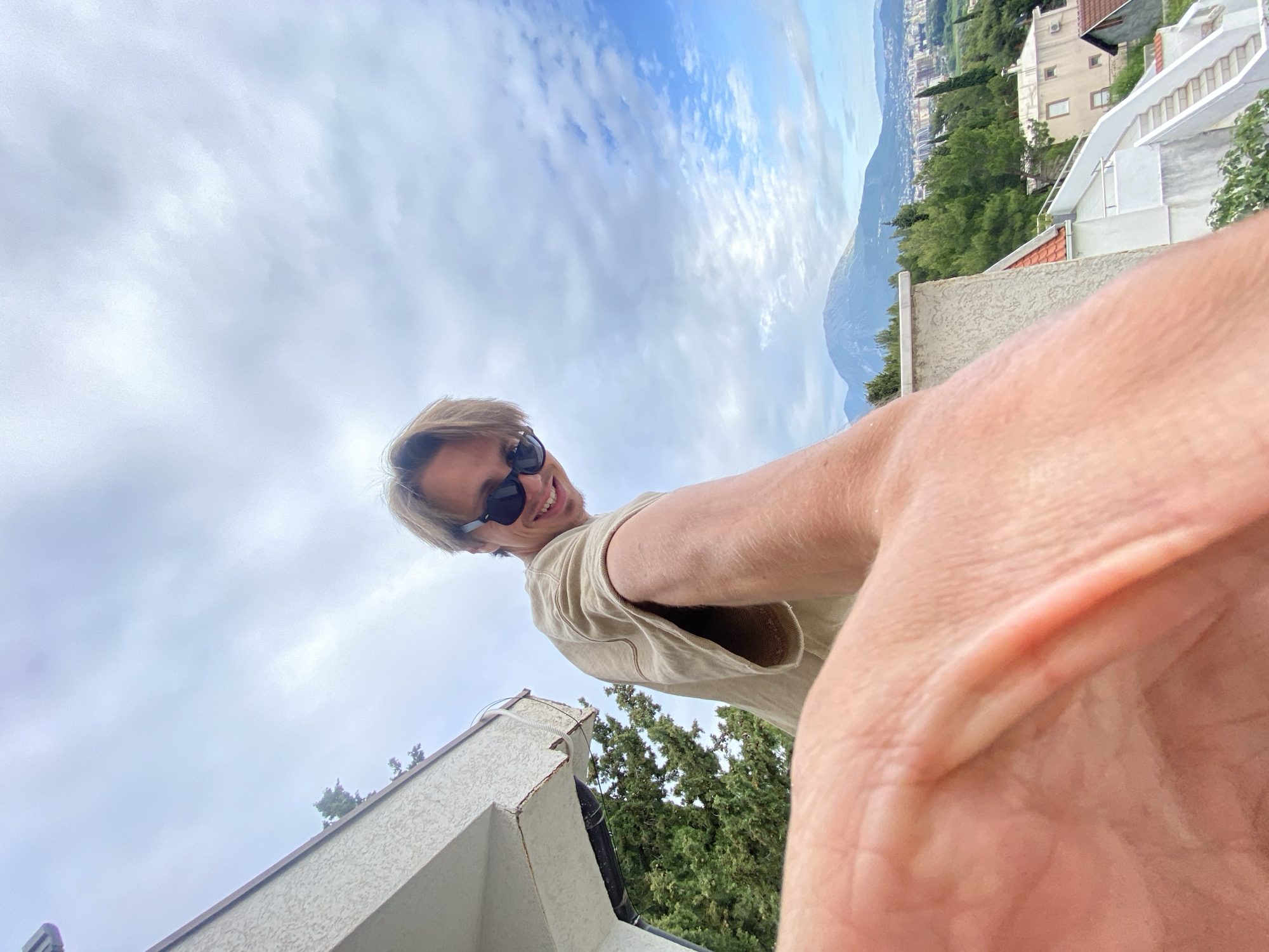 A selfie taken at a low angle, shows a man with short blonde hair and sunglasses, wearing a light tan shirt. In the background is a cityscape and mountains under a cloudy sky, appearing to be Šušanj, Montenegro. Part of the man's outstretched arm and hand are prominently in the foreground, obscuring the view. Elements of buildings and trees are also visible around the man.