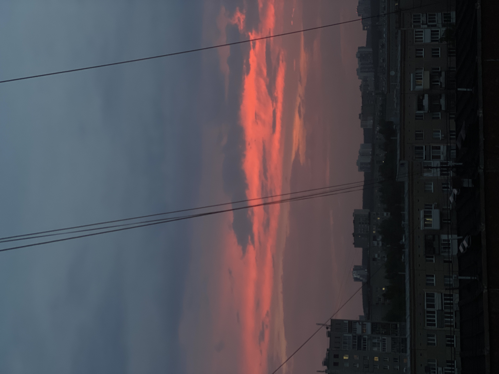 The image depicts a dusky sky over a city skyline in Izhevsk, Russia. The sky is a dramatic gradient of deep blue at the top transitioning to a vibrant reddish-pink hue at the horizon, illuminated by the setting sun. Clouds are scattered across the sky, catching the red light and adding texture.
In the foreground and midground, the silhouette of an urban landscape is visible. Buildings of varying heights and architectural styles are clustered together, appearing as dark shapes against the colorful sky. Several tall apartment buildings, characteristic of Soviet-era urban planning, are prominent. Their windows, some illuminated with yellow light, suggest occupancy. Power lines, taut and thin, crisscross the frame, dividing the sky and drawing the eye across the scene. The overall impression is one of urban life settling down for the night under a strikingly colorful twilight sky.