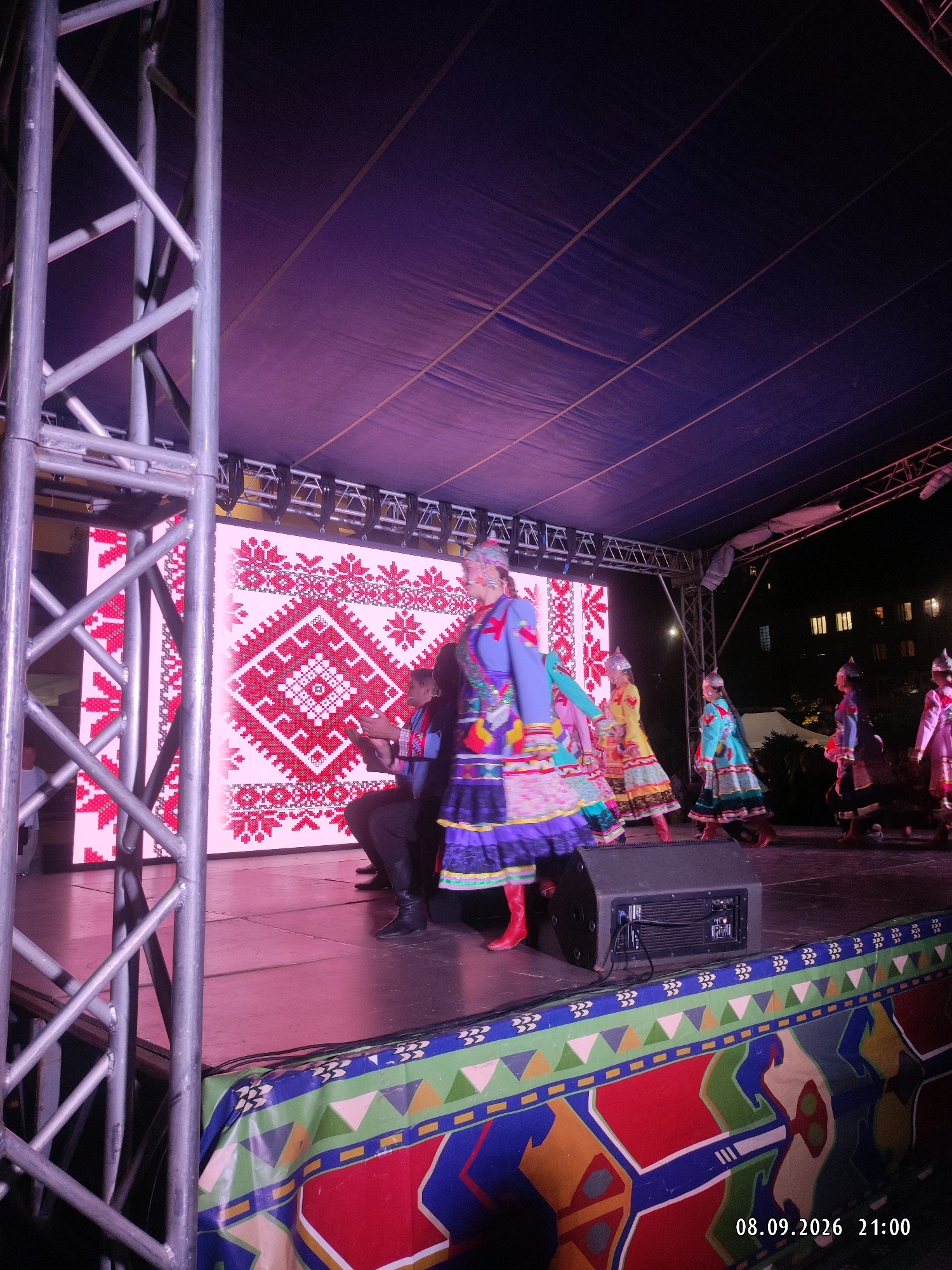 A staged outdoor performance is captured at night, under a large purple tarp. In the foreground, a metal truss structure stands on the left, and a colorful decorative banner with geometric patterns spans the bottom of the image. The stage floor is a dark surface.
On the stage, a group of people are performing. Most are wearing vibrant, traditional costumes with intricate embroidery and colorful skirts. The costumes feature red boots. One performer in the center wears a blue and purple jacket over a multicolored skirt, and a headpiece. Another performer in a yellow dress is to her right, and other performers in blue and pink costumes are visible further back. A man in a dark suit and hat is seated on the stage to the left, holding what appears to be a musical instrument.
Behind the performers, a large LED screen displays a pink and white traditional geometric pattern. To the right of the stage, out of focus, are some trees and buildings with illuminated windows, suggesting an urban environment. In the bottom right corner, a date stamp reads "08.09.2026 21:00".
The image does not provide any specific details that would pinpoint the location within the city of Makhachkala, Russia.