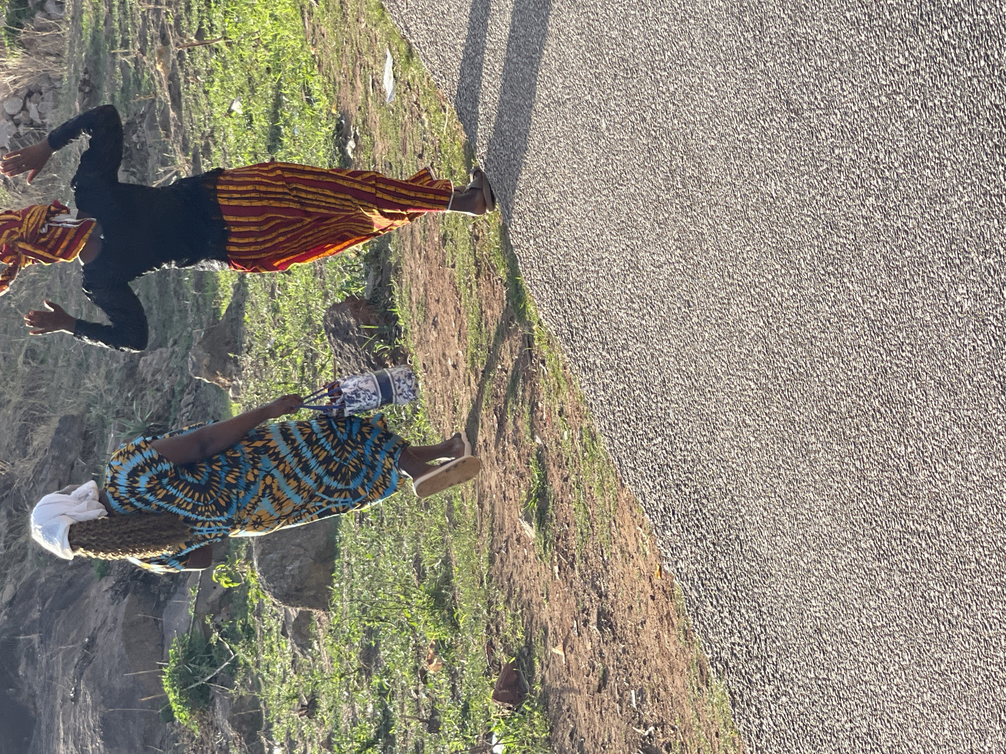 Two women walk along a road in Jos, Nigeria. One woman wears a black long-sleeved shirt and a red and yellow striped skirt, with a matching head wrap. She extends both arms out to the side. The other woman wears a blue, gold, and black patterned dress, and a white head wrap. She carries a purse and wears sandals. The women are walking on the grassy edge of a paved road, with shadows cast on the road. Grass and some rock formations line the adjacent terrain.