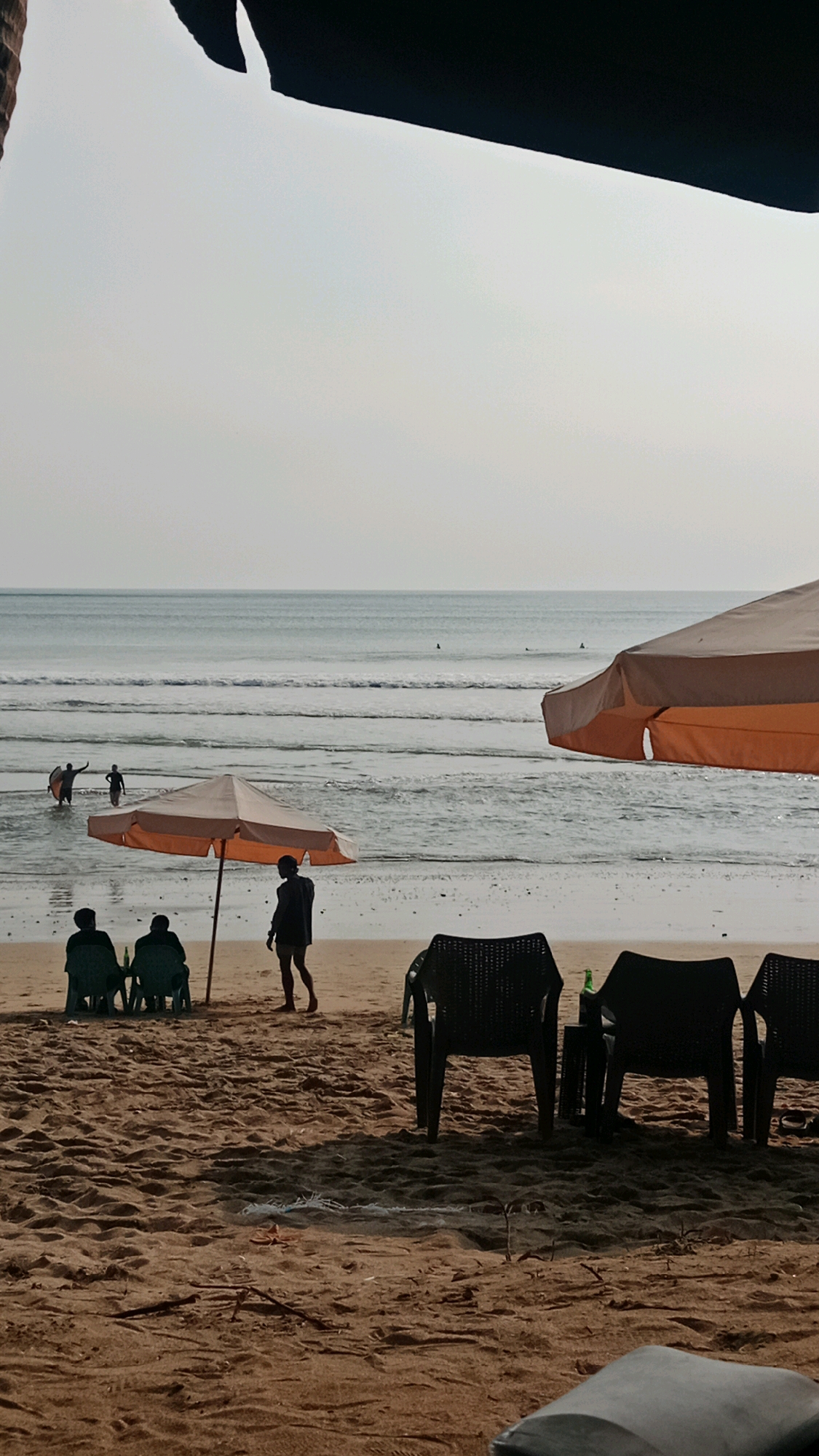 This is a photograph taken from a beach in Kuta, Indonesia, likely during the late afternoon or early evening. The perspective is from slightly above eye level, looking towards the ocean. In the foreground, there is a sandy beach with several black plastic chairs and a small table with a green bottle on it. Two large, orange beach umbrellas are visible; one is in the middle ground, angled slightly to the left, and the other is in the right foreground, partially obscuring the view.
Further back, on the wet sand near the water's edge, a solitary figure stands facing the ocean. To the left of this figure, two people are seated on green chairs under the middle umbrella. In the water, several individuals are visible, some appearing to be surfers or swimmers. The ocean is calm with gentle waves rolling in, and the sky is overcast, suggesting a soft, diffused light. The overall impression is one of a relaxed beach scene.