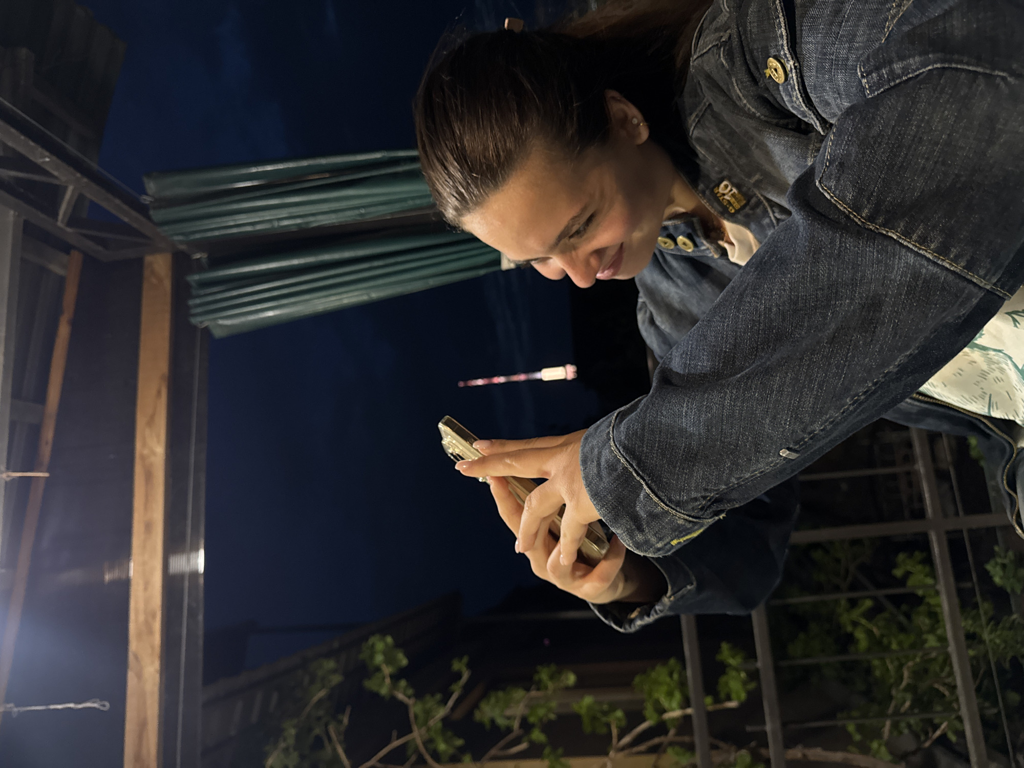 A woman is looking down at her smartphone, which she is holding in both hands, in Yerevan, Armenia at night. She is wearing a denim jacket and standing outside, with a metal fence and tree branches visible in the lower right corner of the image. There is a structure with corrugated sheets overhead and a wooden post on the left side. A light is visible in the distance.