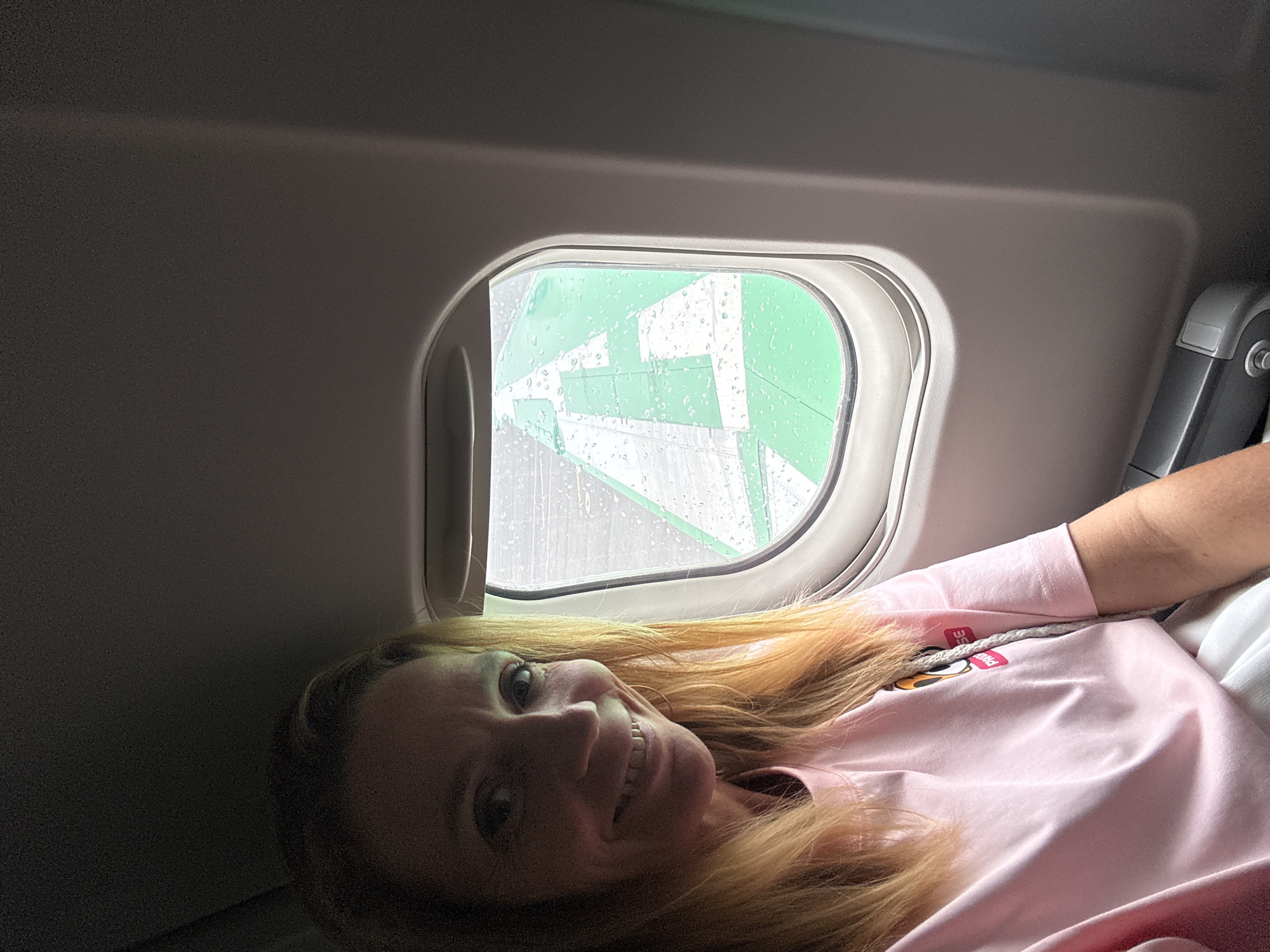 A woman is lying by an airplane window. Outside the window, it is raining on the wing of an Uzbekistan Airways plane. This photo was likely taken at the Tashkent International Airport (TAS) in Tashkent, Uzbekistan.