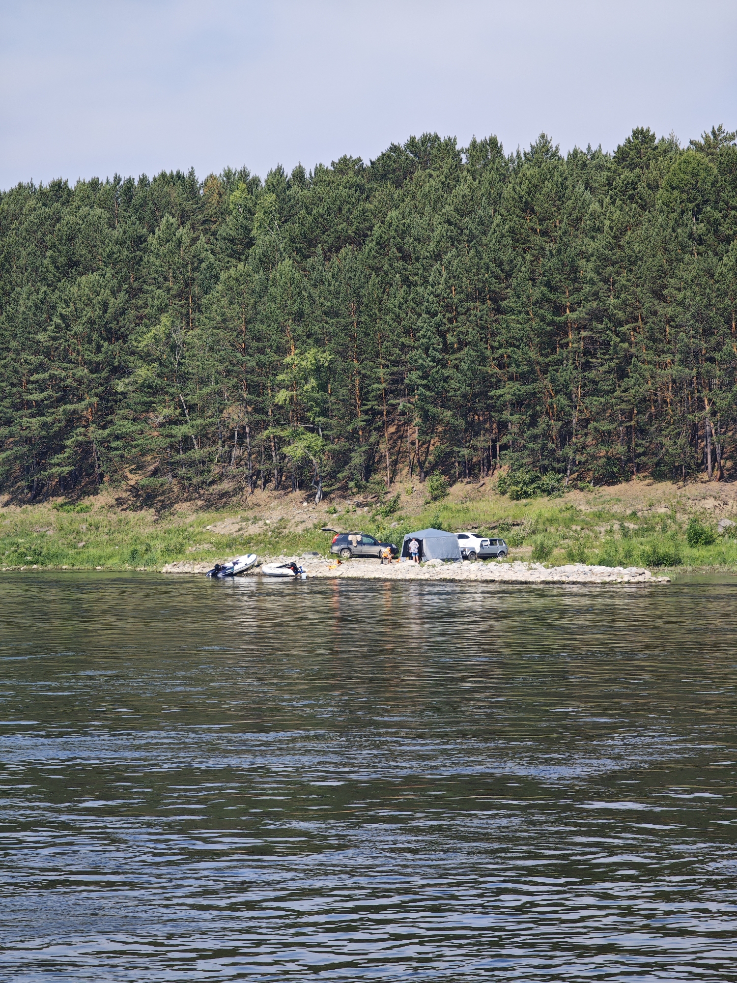 A campsite is set up on a rocky shore of a wide, calm river. A grey tent is pitched on the gravel, with two inflatable boats, one white and one grey, pulled up onto the shore beside it. Two cars, a dark grey SUV with its trunk open and a lighter grey SUV, are parked near the tent. A person stands by the tent, and another person is visible near the dark grey SUV. The river reflects the blue sky and the dense green pine forest that lines the opposite bank. The forest covers a gently sloping hillside, with some dry grass and scrub brush visible in the foreground of the bank. The sky is mostly clear with a few wispy clouds. The scene appears to be a remote camping spot, possibly in the rural area surrounding Podporozh'e, Russia, given its natural landscape.