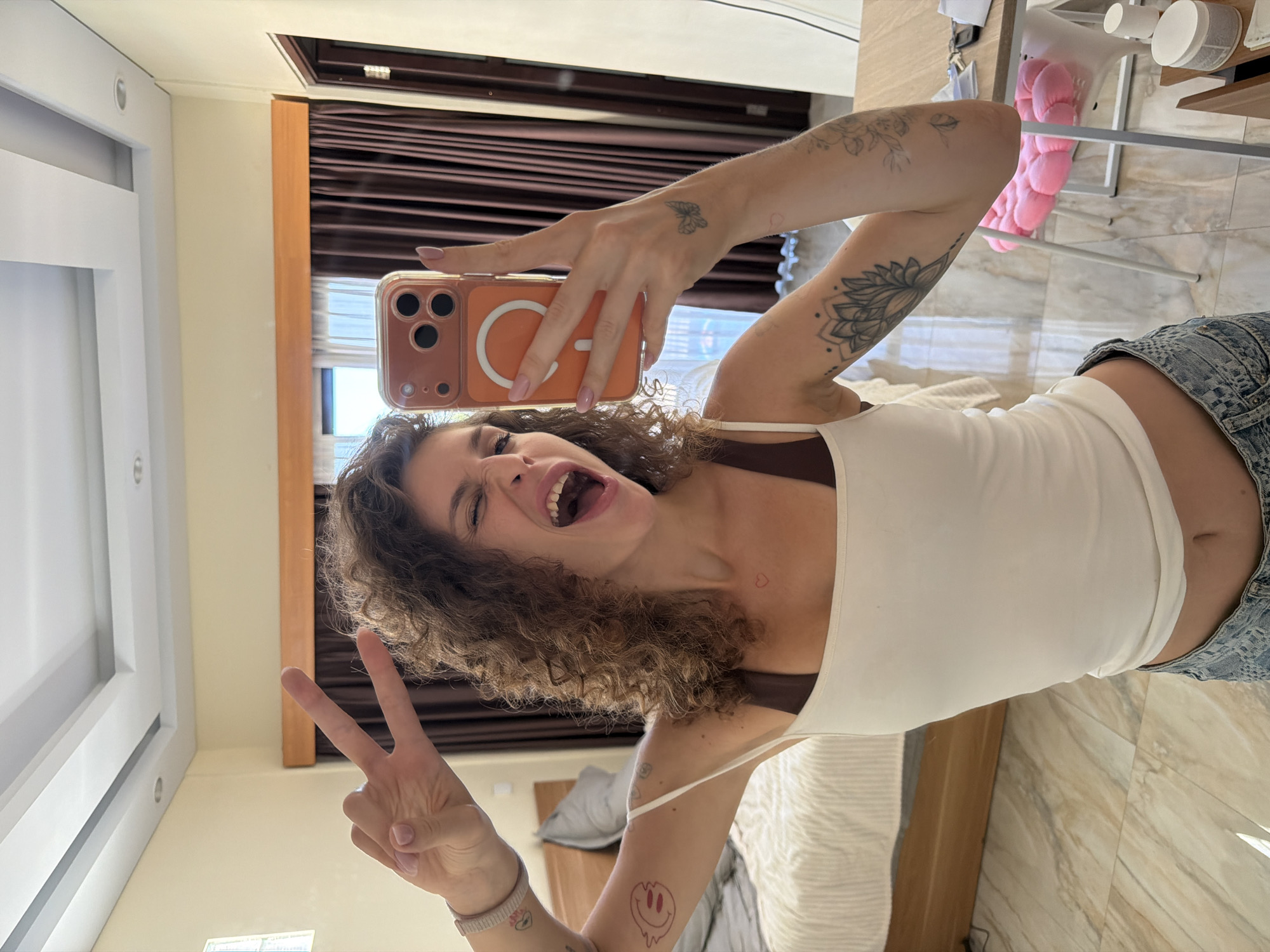 A woman is taking a selfie indoors, likely in a hotel room in North Kuta, Indonesia. She is wearing a white tank top and denim shorts, has curly hair, and is holding up a peace sign with her left hand. Her phone, encased in an orange case, obscures part of her face. She has tattoos on her arms and chest. The background reveals a room with brown curtains, a bed, and a white chair. The floor is light-colored tile.