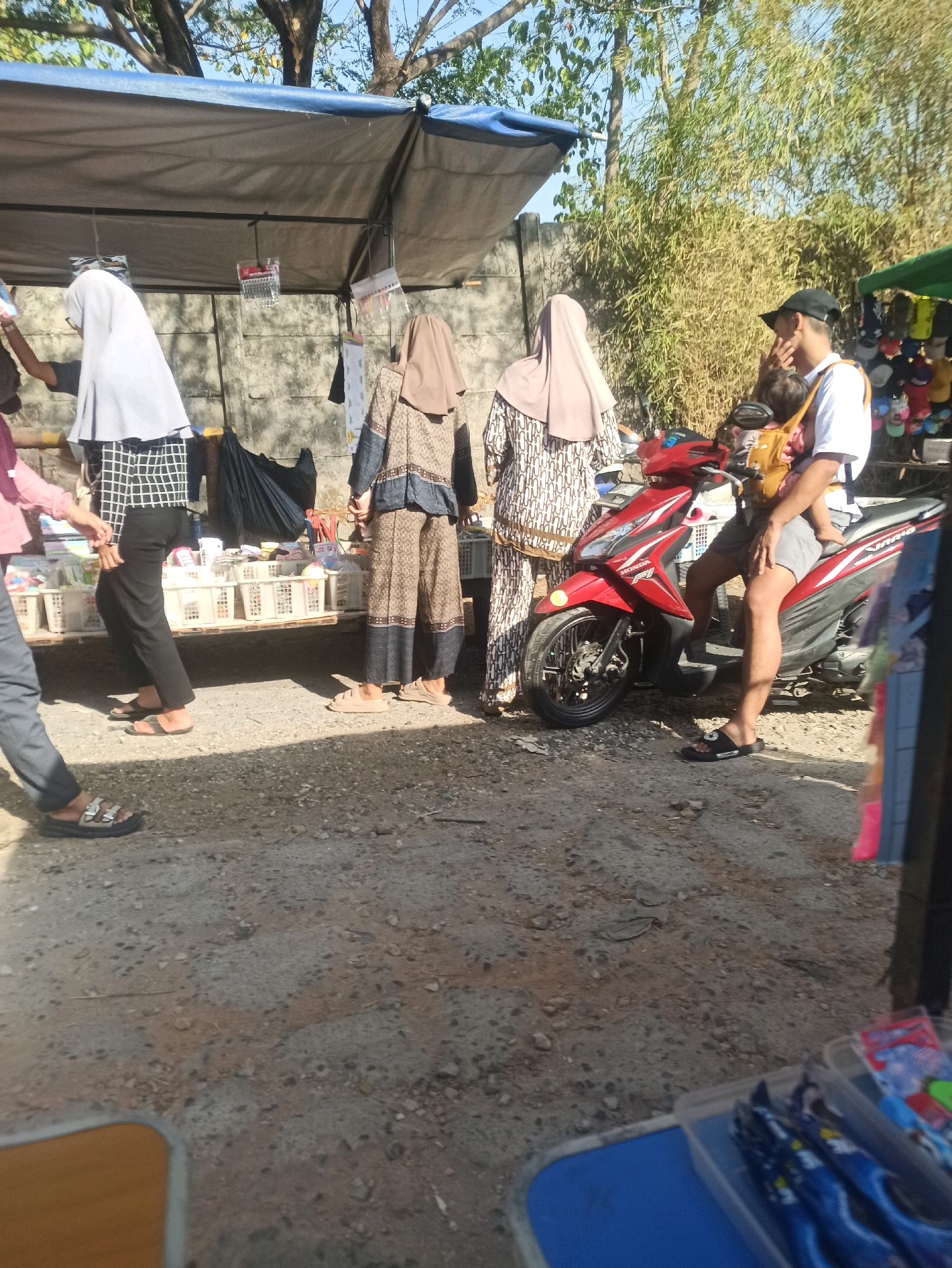 The image depicts a street scene in Telagamurni, Indonesia, likely a marketplace, under a clear sky with visible trees and bamboo. A red Honda motorcycle is parked in the foreground, with a man wearing a white t-shirt, grey shorts, a black cap, and a baby carrier with a child in it. To the left of the motorcycle, two women wearing hijabs and traditional Indonesian clothing stand near a stall displaying various items in white baskets. One woman wears a white hijab and a patterned black and white outfit, while the other wears a light brown hijab and a patterned brown and beige dress. Another person in dark pants and sandals is partially visible on the far left, appearing to be browsing. The ground is a mix of dirt and gravel. In the background, a shaded tent covers the stall, and further back, more stalls with merchandise like hats are visible.