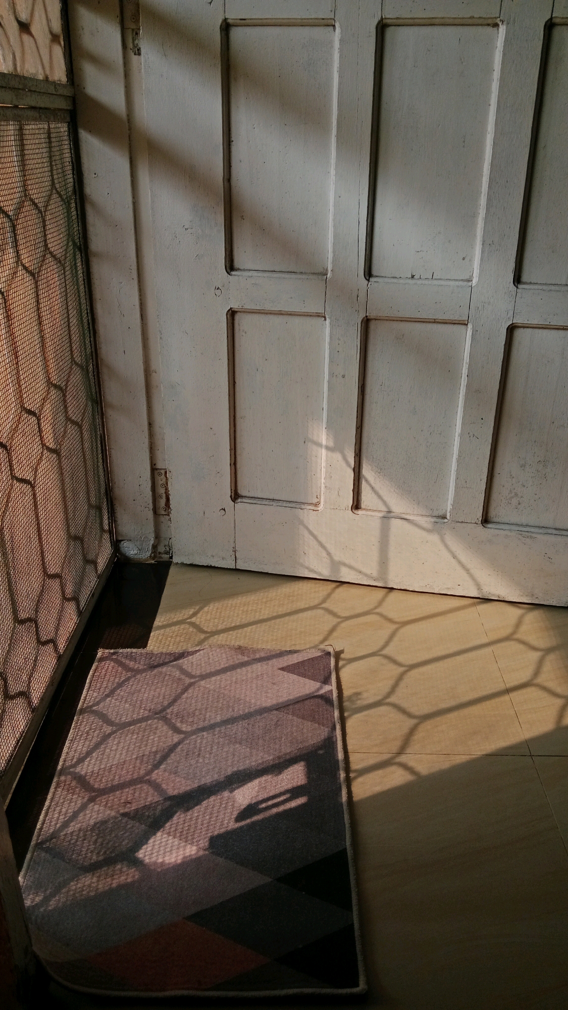 The image shows a close-up view of a doorway and a doormat, bathed in sunlight that casts geometric shadows. The doorway is made of white painted wood with four recessed panels. To the left of the doorway is a screen door with a hexagonal mesh pattern. A doormat with a diamond-shaped pattern in shades of gray, brown, and pink is placed in front of the doorway. The floor appears to be tiled and is also covered in the same geometric shadows. The scene suggests an indoor or covered outdoor space, possibly a home or building in Tanjung Pinang, Indonesia, given the sunlight patterns often seen in tropical regions. No people are visible in the image, and no specific actions or interactions are depicted.