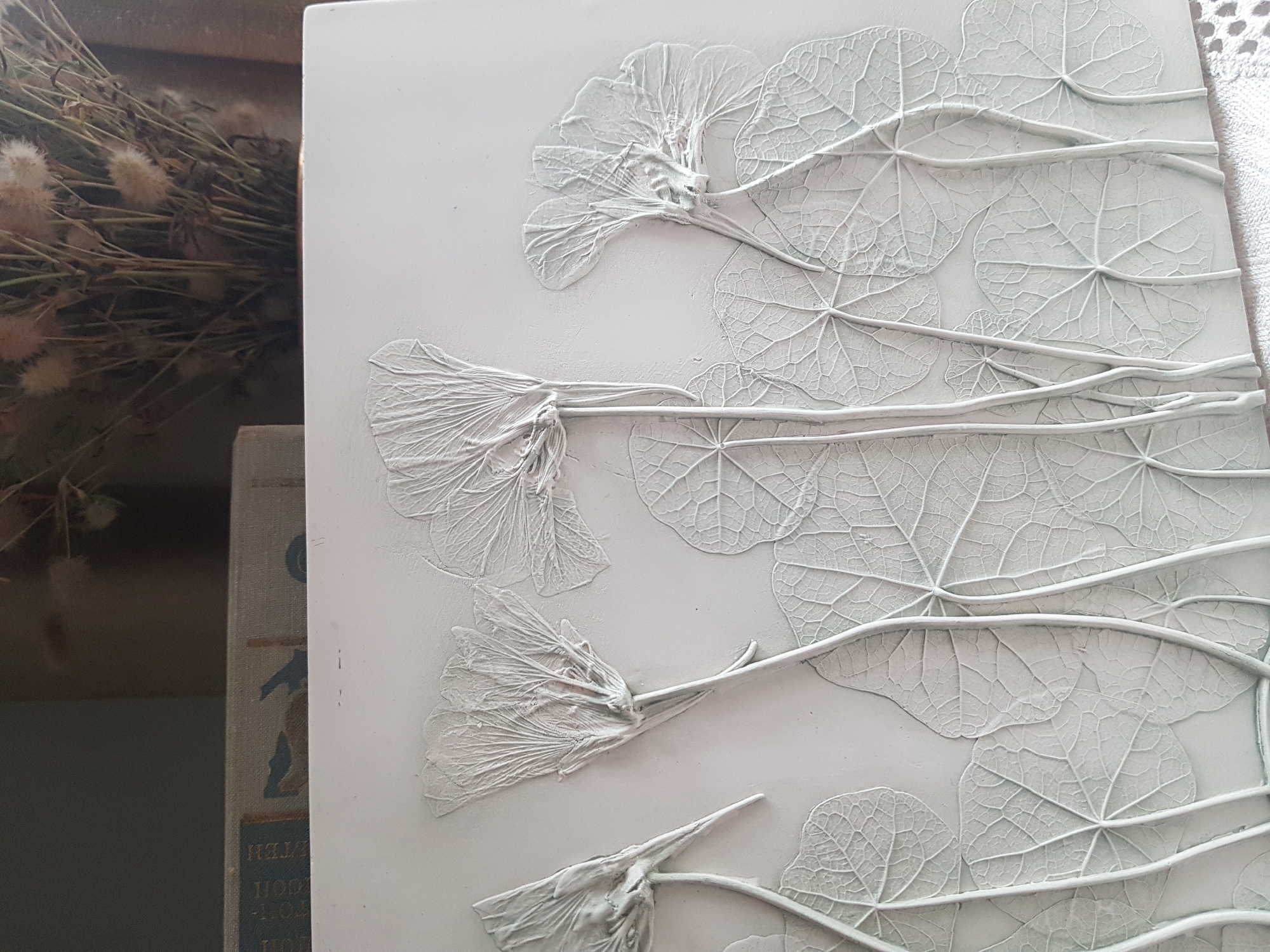 A high-angle close-up shot presents a white, square artwork featuring a raised design of flowers and leaves. The flowers, characterized by their delicate, vein-like petals, appear at various angles, with some positioned at the top and others towards the bottom of the frame. Interspersed among the flowers are round leaves with prominent veins, adding texture and depth to the composition. The artwork is predominantly white, with subtle variations in tone highlighting the raised elements. To the left, a stack of books is visible, with the spine of the topmost book displaying worn blue fabric and gold lettering. Adjacent to the books, a bouquet of dried flowers adds a rustic touch to the scene. The setting is suggestive of an indoor environment, potentially an artist's studio or a display in Saint Petersburg, Russia.