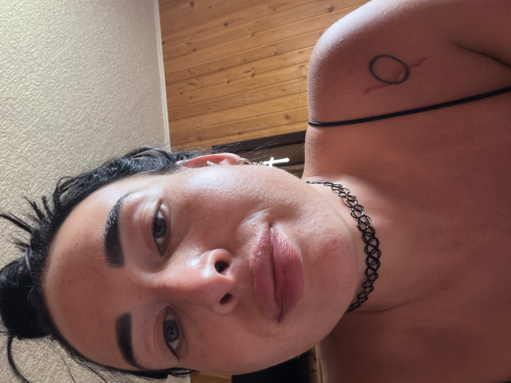A close-up, low-angle shot shows a woman's face from the chest up, against a background of wood-paneled walls and a textured ceiling. Her dark hair is pulled up into a messy bun, with some strands falling around her face. She wears a black choker necklace and a thin black strap across her shoulder, from which a cross-shaped earring dangles. A small tattoo of a circle with a line through it is visible on her right shoulder. The woman has dark eyebrows, dark eyes, and her lips are slightly parted. The lighting suggests an indoor environment, possibly a home. The location is identified as Sochi, Russia.