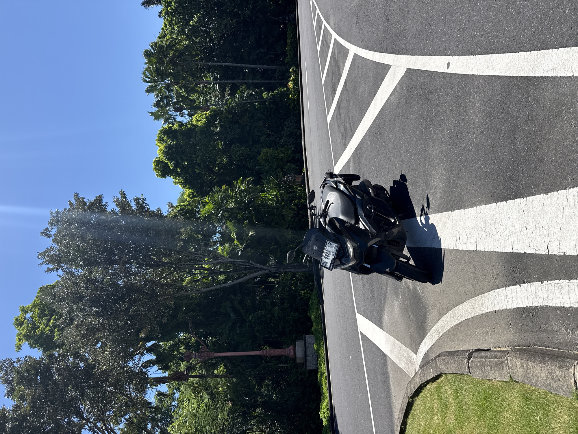 A motorcycle is parked on the side of a road in Nusa Dua, Indonesia. The motorcycle is black and is parked in a designated parking area marked by white lines on the asphalt. A curb separates the road from a grassy area, and a red ornate lamppost stands near a cluster of trees and greenery. The sky is blue, and the vegetation includes palm trees and other tropical plants. The motorcycle's license plate is partially visible.