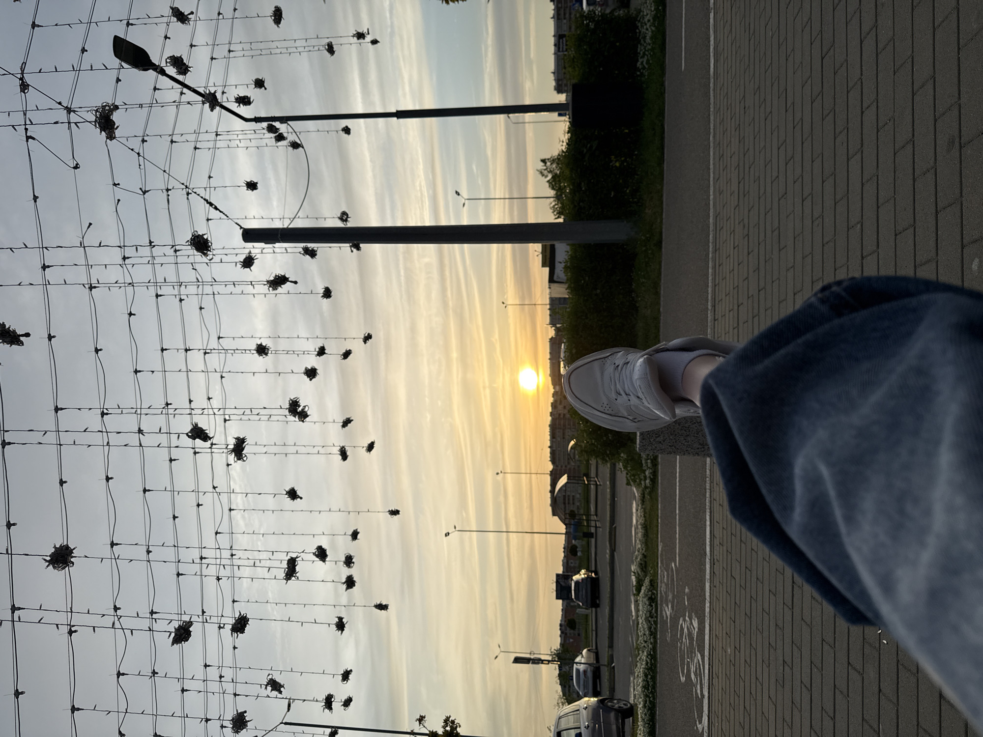The image captures a sunset view from a city location, focusing on a first-person perspective. A leg with a white sneaker and blue jeans is visible in the lower right. The background features a street with parked cars, street lights, and buildings silhouetted against the sunset. Overhead, there is a grid-like structure with decorative elements. The sky is filled with yellow and orange hues from the setting sun. The city is unnamed in the image.