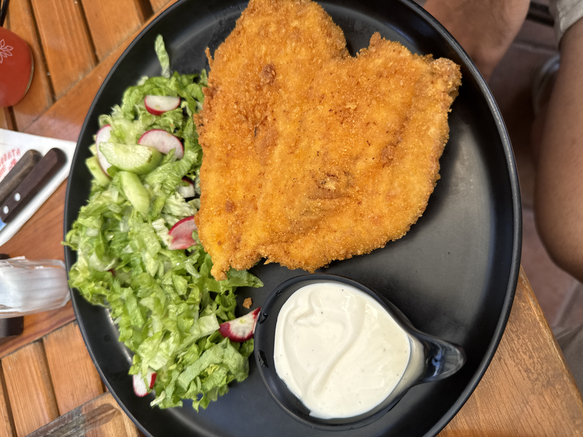 A close-up, overhead shot captures a meal served on a black, circular plate resting on a wooden table. The plate contains a large, golden-brown, breaded cutlet, likely chicken or pork, positioned to the right. To the left of the cutlet, a small black dish holds a generous dollop of white sauce, possibly ranch or aioli. Further left and slightly above, a salad composed of chopped green lettuce, thin slices of radish, and cucumber rounds is presented. The wooden table's grain is visible, suggesting an outdoor or rustic dining setting. In the background, out of focus, a clear glass with ice and a drink in a red cup are visible, along with part of a napkin and cutlery. The overall scene appears to be a casual meal enjoyed at an outdoor establishment, possibly a restaurant or cafe. Specific details regarding location in Sofia, Bulgaria are not discernible from the image.