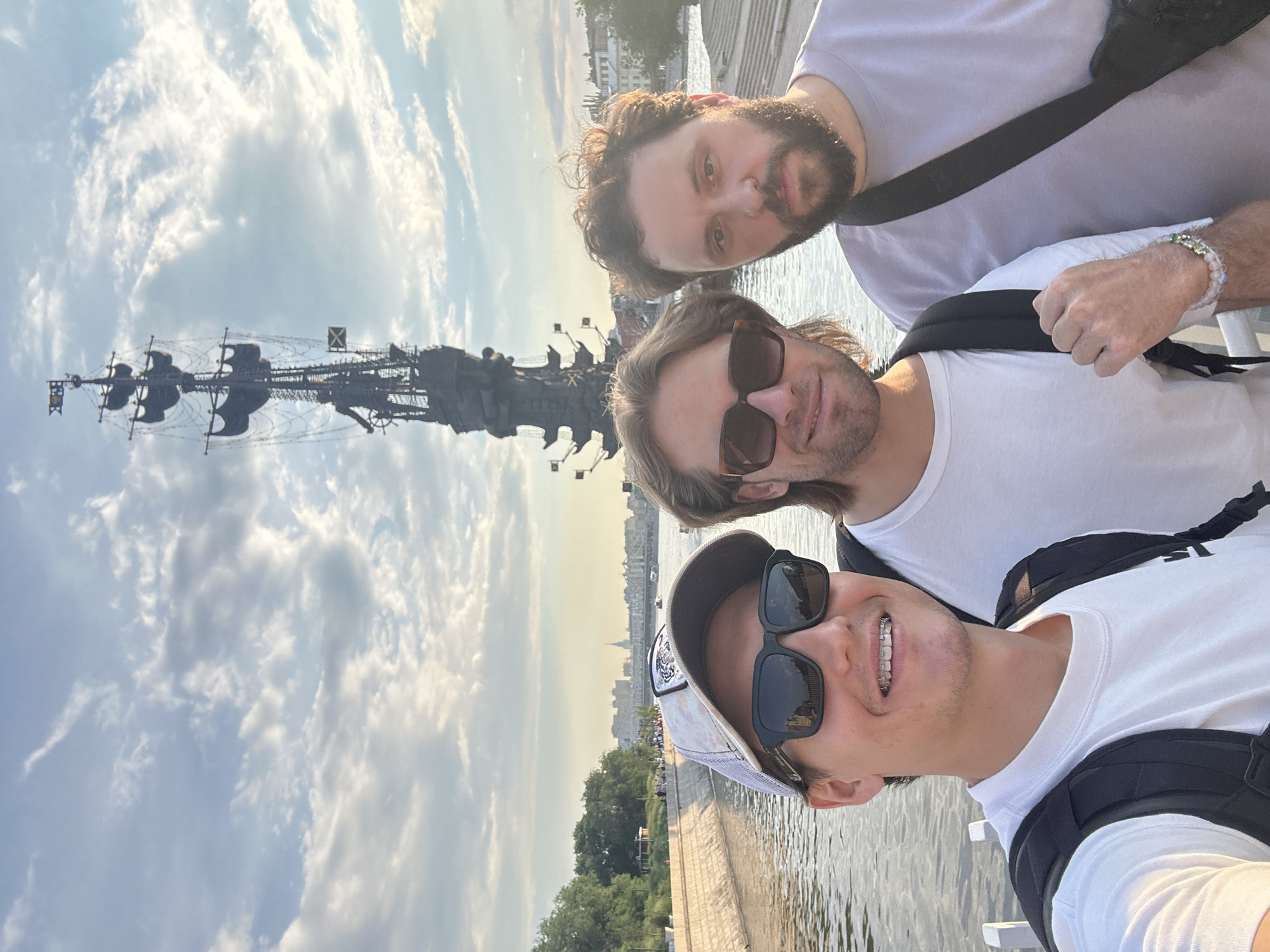 This is a selfie taken outdoors in Moscow, Russia, featuring three men standing in front of the Peter the Great statue on a sunny day with scattered clouds. The statue, a massive monument depicting Peter the Great on a ship, dominates the background. The men are positioned in the foreground, with the man on the left in a white t-shirt, a baseball cap, and sunglasses, smiling and showing his braces. The middle man wears a white t-shirt and sunglasses, while the man on the right is in a light purple t-shirt and has a beard. They are all wearing backpacks, suggesting they are tourists. The environment includes a body of water, likely a river or canal, with a stone embankment and trees on the far side. In the distance, the Moscow skyline is visible, with buildings and what appears to be a bridge.