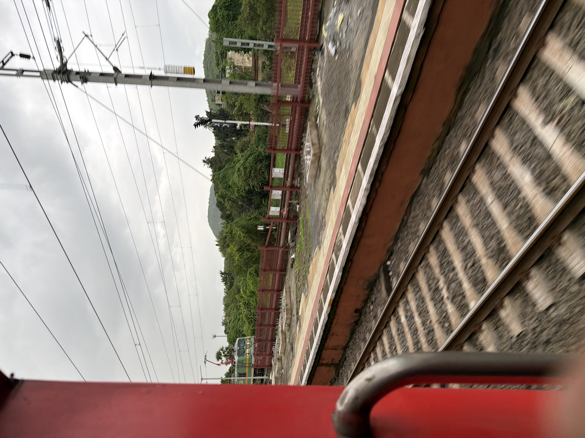 A train moves past a station on a cloudy day. The perspective is from within the train, looking out a window. A portion of a red train car is visible on the left, obscuring a portion of the view. In the distance, a green and white train engine is stopped on the tracks. A red fence lines the platform of the station. Overhead, power lines crisscross the sky. Lush green trees and hills form the background.
