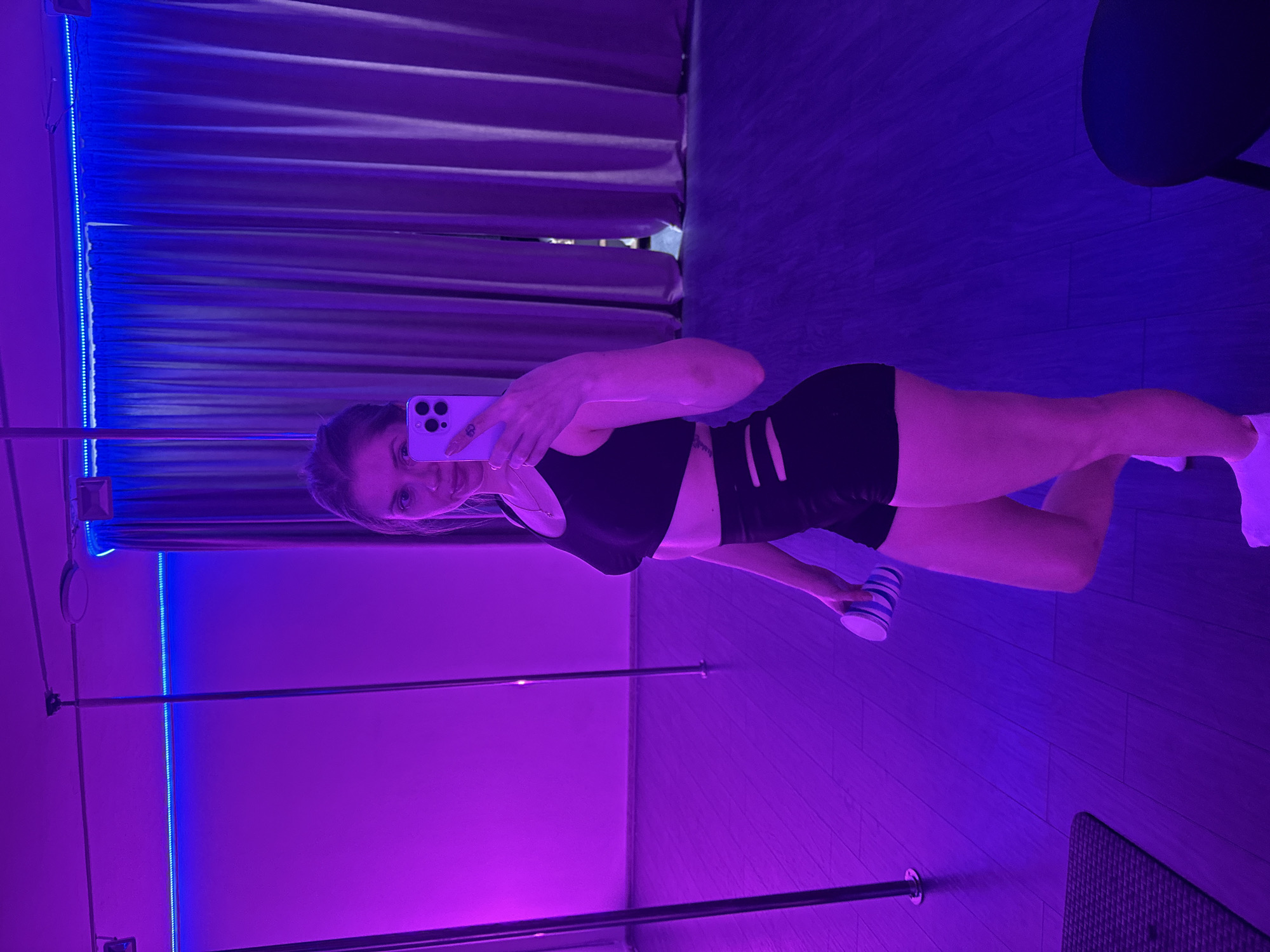 A female is taking a selfie in a studio with a pole. She is wearing a black sports bra and black shorts. She is holding a blue and white striped cup in her right hand. The studio is lit with blue and purple LED lighting. There are curtains behind her. The floor appears to be made of wood or laminate planks. A black stool or table is in the background. The city of Batumi, Georgia is not discernible from the image.