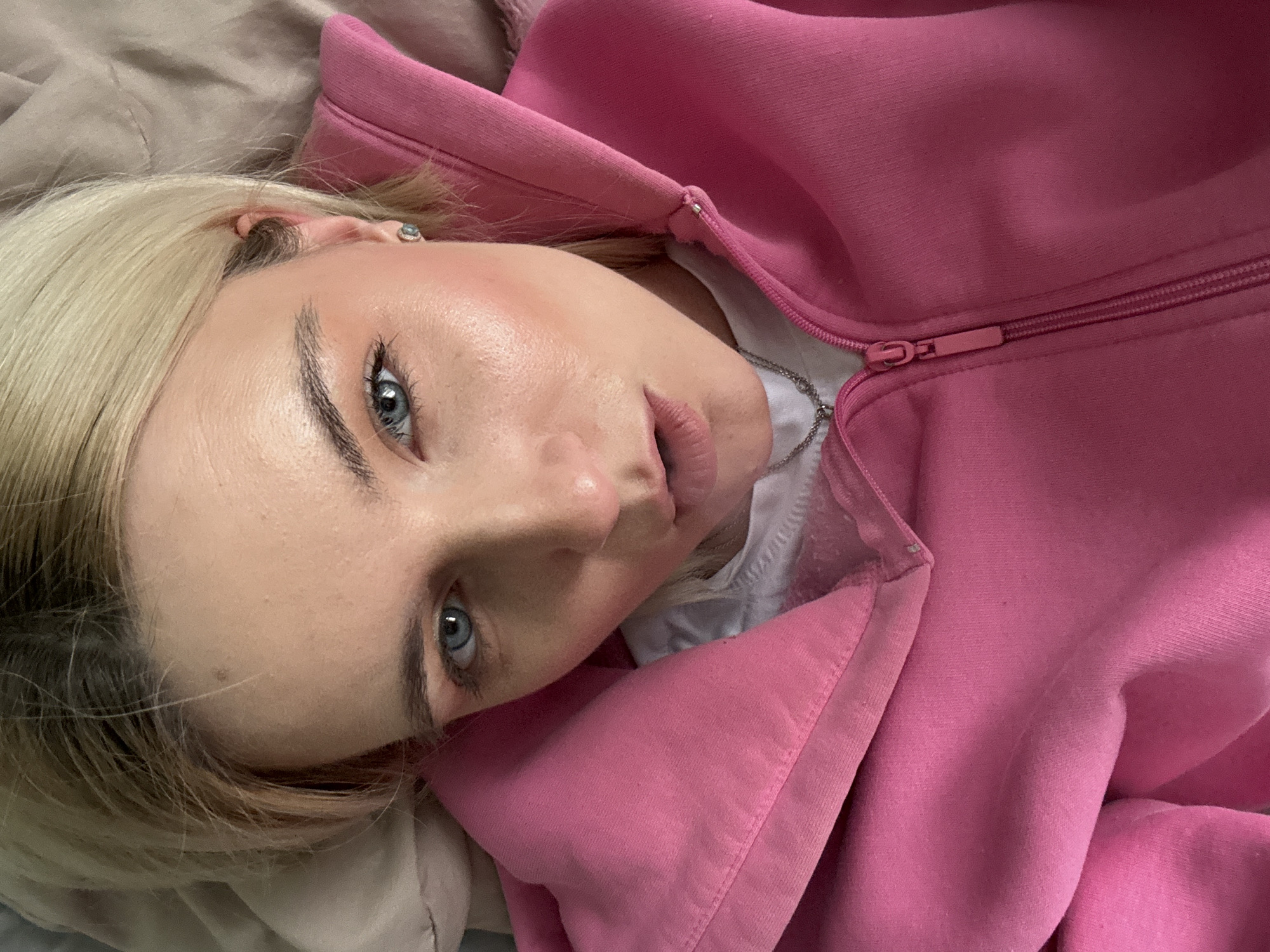 A person with fair skin and light blonde hair is lying down, wearing a pink hooded sweatshirt partially zipped open to reveal a white T-shirt and a silver necklace. The person is positioned at an angle, with their head tilted towards the camera, and their blue eyes are looking directly at the viewer. The background consists of a neutral-toned bedding material. The location is not discernible from the image.