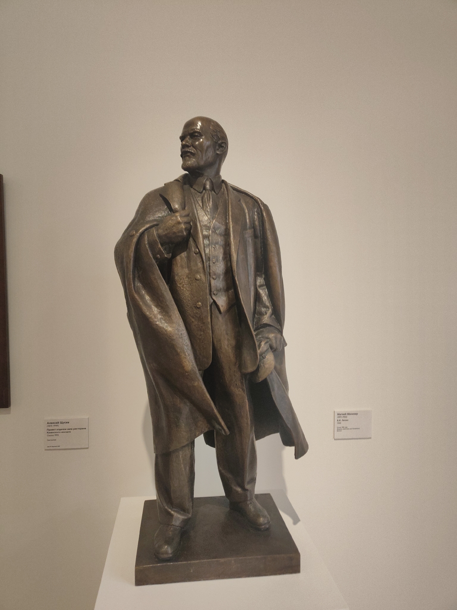 The image shows a bronze statue of Vladimir Lenin, a prominent historical figure, standing on a dark brown square base. Lenin is depicted in a suit, vest, and overcoat draped over his left arm, suggesting a formal or authoritative presence. The statue is set against a plain, light-colored wall in what appears to be a museum or gallery setting, indicated by two information plaques visible on either side of the statue. The plaque to the left identifies "Алексей Щусев" (Alexey Shchusev) (1873-1947) and refers to a project for decorating the restaurant hall of the Kazan train station around 1915. The plaque to the right identifies "Матвей Манизер" (Matvey Manizer) (1891-1966) and references "В.И. Ленин" (V.I. Lenin) in 1940. The details around the statue are not particularly clear, however, it appears to be located in the city of Unknown, Unknown. The neutral background and straightforward presentation of the statue suggest a setting where artifacts or historical figures are displayed for public viewing or educational purposes.