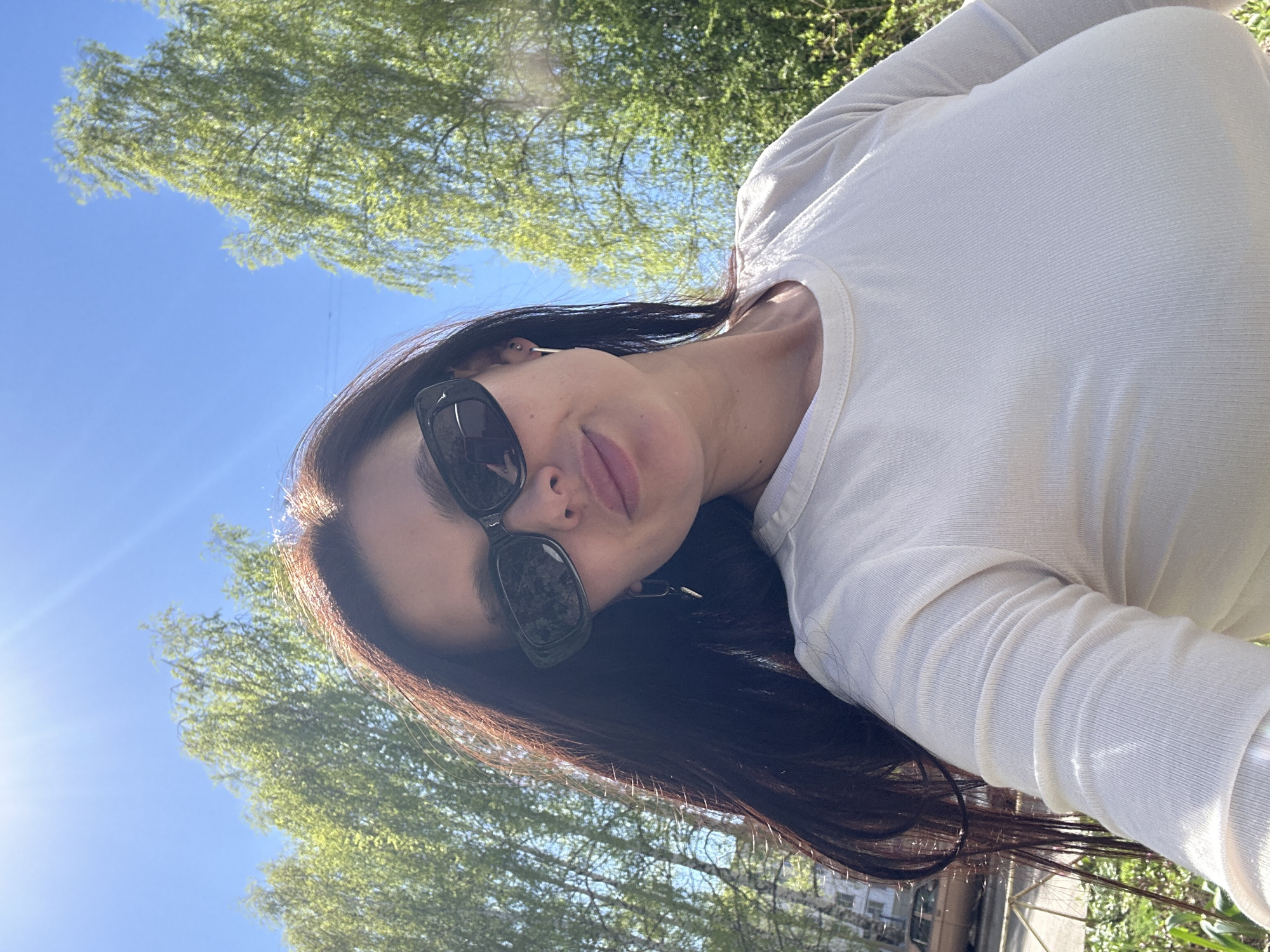 A young woman is outdoors, likely in the city of Chaikovsky, Russia. She is wearing sunglasses and a long-sleeved white top. She is standing outside, with trees and a blue sky with sunlight visible in the background. The presence of trees and buildings in the background suggests a park or urban green space. The photo appears to be taken at an angle, possibly as a selfie.