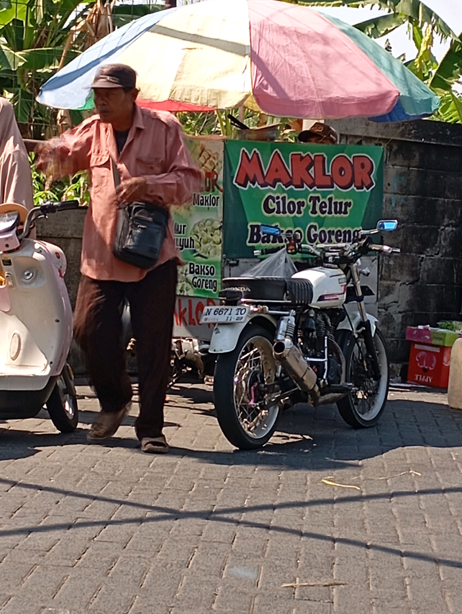 In Kalisampurno, Indonesia, a man wearing a brown cap and a light salmon-colored long-sleeved shirt stands in front of a food stall. He has a dark-colored shoulder bag. Beside him, to the left, is a light-colored scooter. To his right and behind him, a white motorcycle is parked. The food stall has a green banner with red lettering that reads "MAKLOR Cilor Telur Bakso Goreng." The man appears to be interacting with someone off-camera or gesturing. The ground is paved with interlocking bricks. In the background, there is a brick wall and foliage. Sunlight casts shadows on the ground.