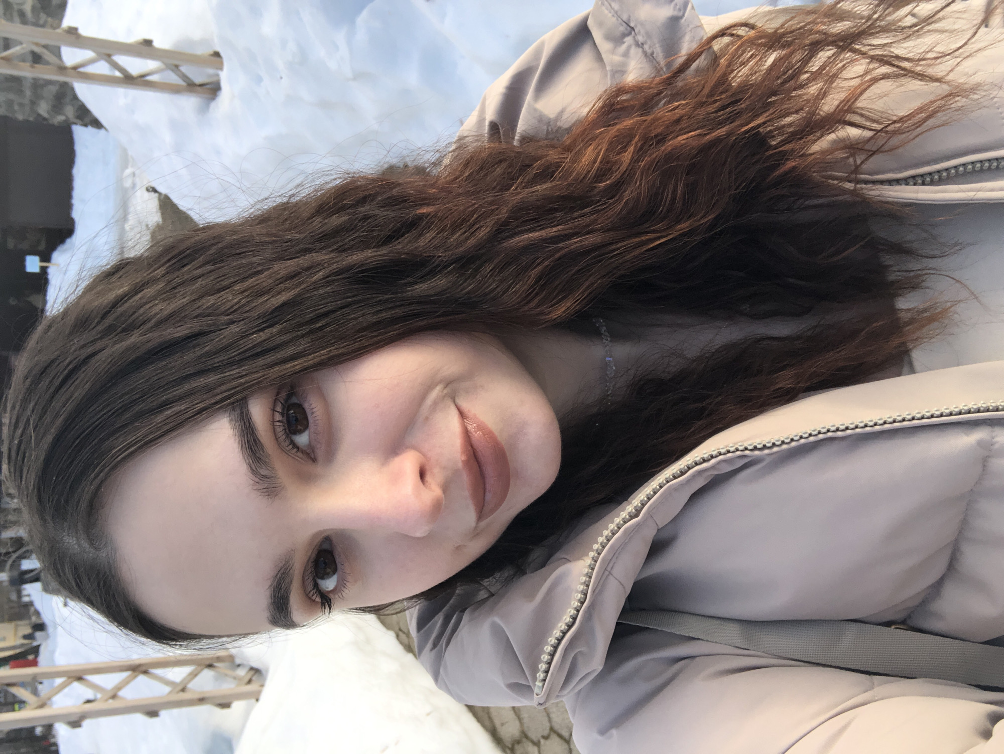 A close-up portrait of a young adult female is captured outdoors in Chaikovsky, Russia, during winter. The subject, centrally framed, possesses long, wavy dark brown hair with visible reddish highlights, dark brown eyes, and a light complexion, enhanced with subtle makeup including light brown lipstick. She wears a light beige or grey padded winter jacket with a central zipper and a visible light grey strap over her right shoulder, along with a delicate necklace. She looks directly at the camera with a slight smile. The background features prominent snowdrifts covering the ground. Visible elements include two light-colored wooden lattice-like structures to her left and right, a dark, stone-like building partially obscured by snow, and a paved or cobbled pathway. A yellow structure and a small blue sign on a pole are faintly discernible in the distant left. The sky appears light blue with diffuse cloud cover.