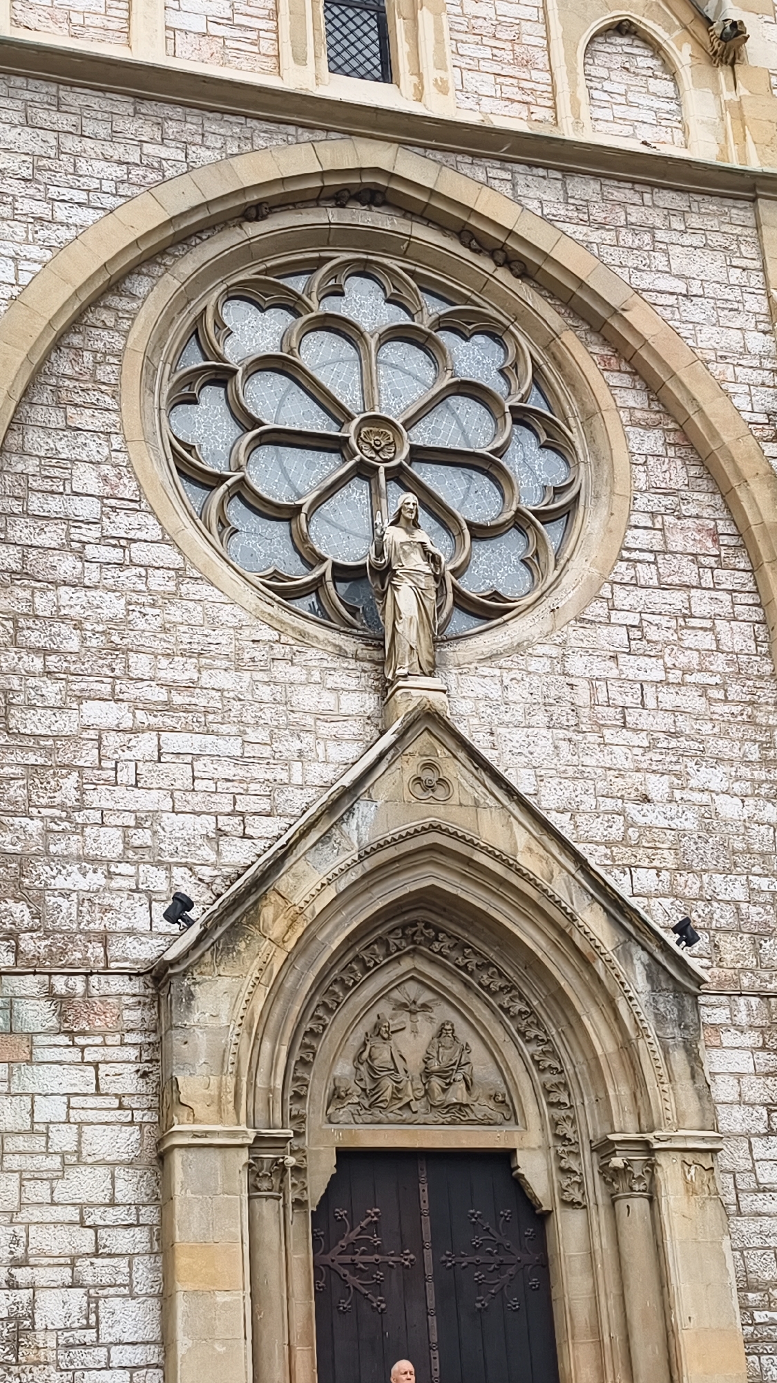 The image shows the facade of a church in Sarajevo, Bosnia and Herzegovina, specifically Saint Joseph's Church. The facade is constructed from light-colored stone blocks arranged in a regular pattern. Dominating the upper portion of the facade is a large, ornate rose window with intricate stone tracery. Below the rose window, a statue of a standing figure, possibly Jesus Christ, is positioned on a small pedestal. This statue is made of a light-colored material and appears to be a religious icon.
The lower part of the facade features a prominent arched entrance with detailed carvings above the dark wooden doors. The carvings within the arch depict two figures seated, with a smaller figure or symbol above them, suggesting a biblical scene. The archway itself is richly decorated with moldings and decorative elements.
Two black security lights are visible on the left side of the facade, mounted on the stone wall above and to the left of the entrance arch. To the right of the entrance, another similar security light is visible on the upper right portion of the facade.
At the very bottom of the image, a portion of a person's head and shoulders is visible, suggesting they are standing in front of the entrance. The person appears to be an older male with white hair and is looking towards the camera.
The overall impression is one of religious architecture with historical and artistic significance, set within an urban environment.