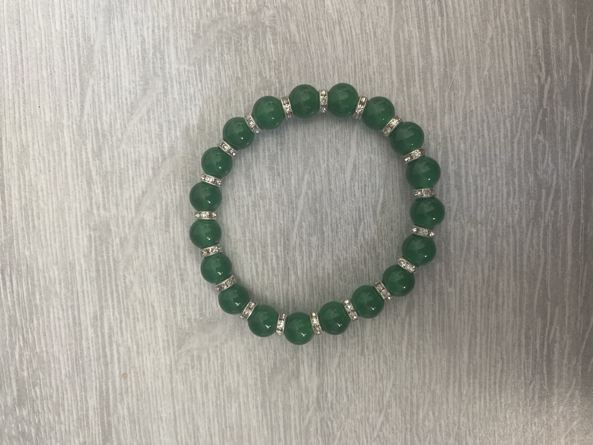 A close-up, top-down, eye-level shot showcases a circular bracelet made of smooth, polished green beads, each separated by a small, silver-toned spacer bead encrusted with clear rhinestones. The bracelet rests on a light gray wooden surface with a visible grain pattern. The lighting is even, casting no harsh shadows. The image is a clear, high-resolution capture, free of any visible dirt, smudges, or distracting elements, suggesting a clean and controlled environment. The scene does not depict any people, actions, or interactions, focusing solely on the static object. The location in Blagoevgrad, Bulgaria, is not visually indicated by any elements within the image.