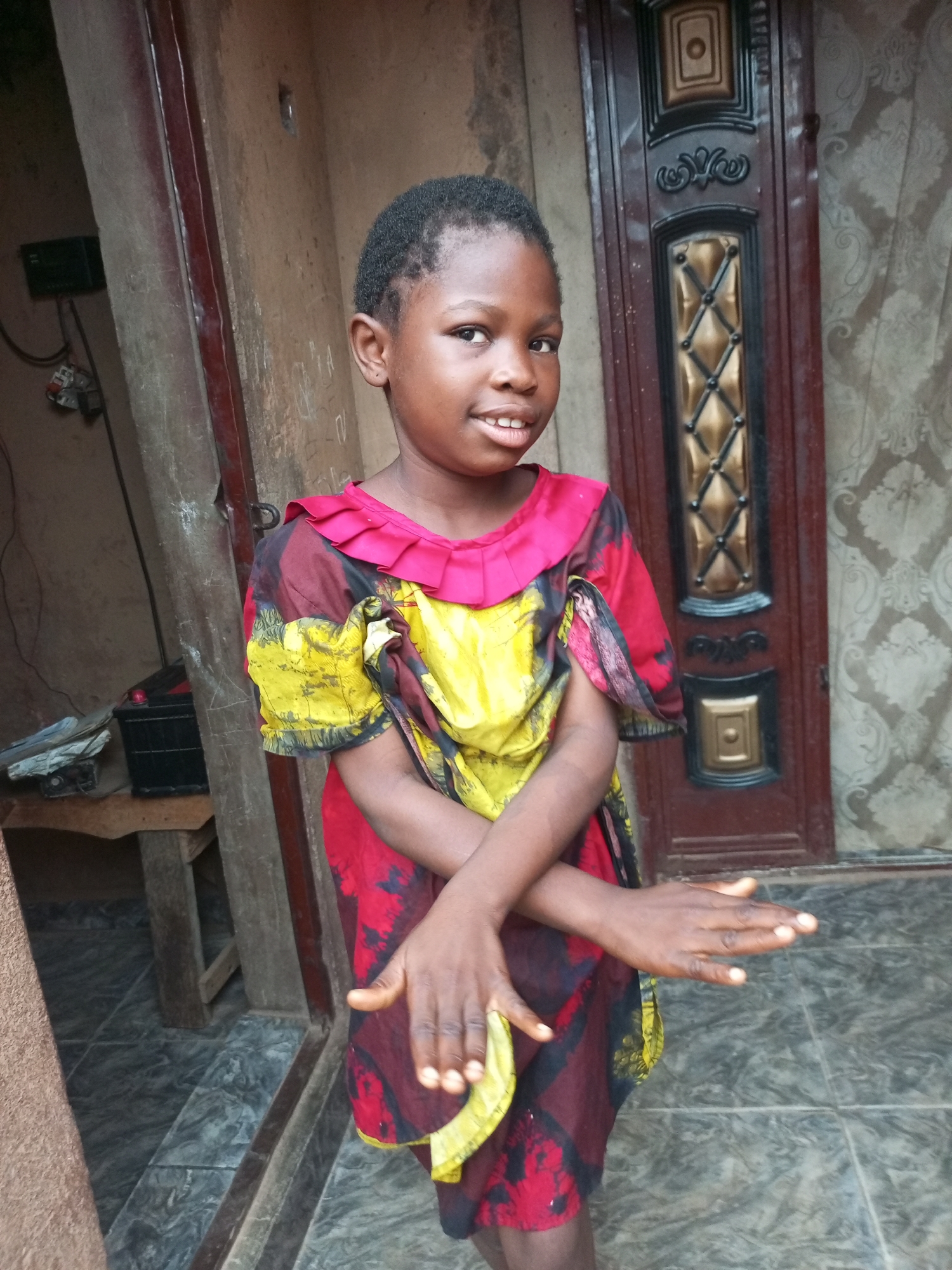 A young girl stands outdoors, facing the camera. She has short, dark hair and is wearing a brightly colored dress with red, yellow, and black patterns. Her hands are crossed in front of her chest, and she has a slight smile. Behind her, there is a dark wooden door with ornate gold and black accents, and a textured wall. To the left of the girl, a wooden table with some objects on it is visible. The ground is tiled. The scene appears to be in a residential area in Tempure, Nigeria.