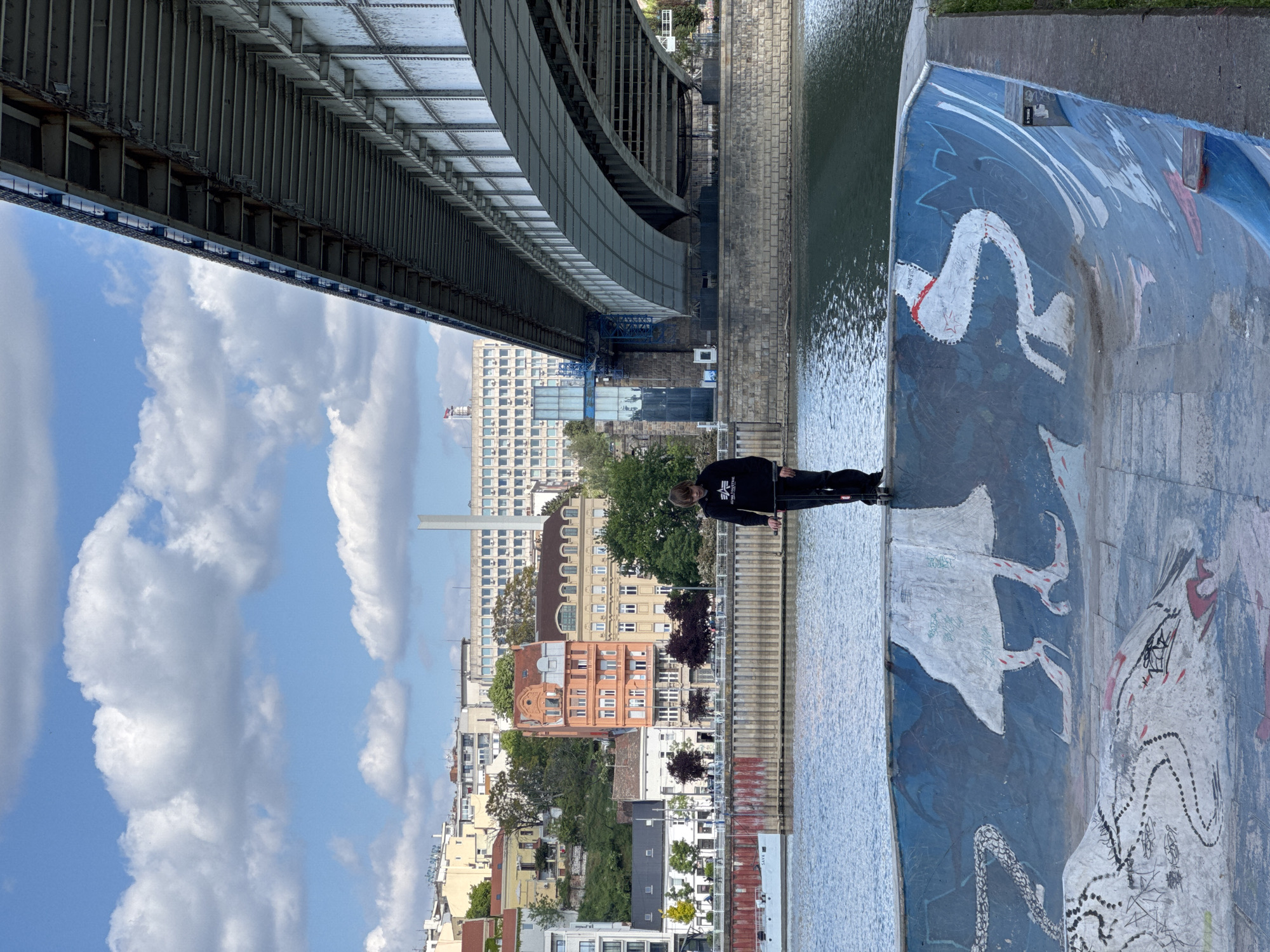The image, when correctly oriented, depicts a daytime scene along a riverbank in Belgrade, Serbia, beneath a large metal bridge. A lone individual, dressed in a dark hoodie displaying a white logo (possibly "ALPHA INDUSTRIES") and dark trousers, stands with their back to the viewer, facing the water.
The upper and left portions of the frame are dominated by the imposing grey metal structure of the bridge, characterized by numerous beams, girders, and riveted panels. The river, calm with gentle ripples and reflections, flows through the mid-ground.
To the right of the person, a concrete embankment or wall curves alongside the river. This surface is decorated with vibrant, stylized murals predominantly in blue, white, and red tones, depicting various figures or creatures, including a long, serpentine form with dotted outlines. Visible text on the painted surface includes "ONELLA B" and "SAVE SAVA," alongside some illegible graffiti.
The background reveals a diverse cityscape under a bright, partly cloudy sky. The buildings showcase a range of architectural styles, from ornate, multi-story structures to more modern, block-like edifices. A tall, slender white tower or chimney rises prominently in the distant skyline. The scene captures a tranquil moment within a busy urban setting during fair weather.