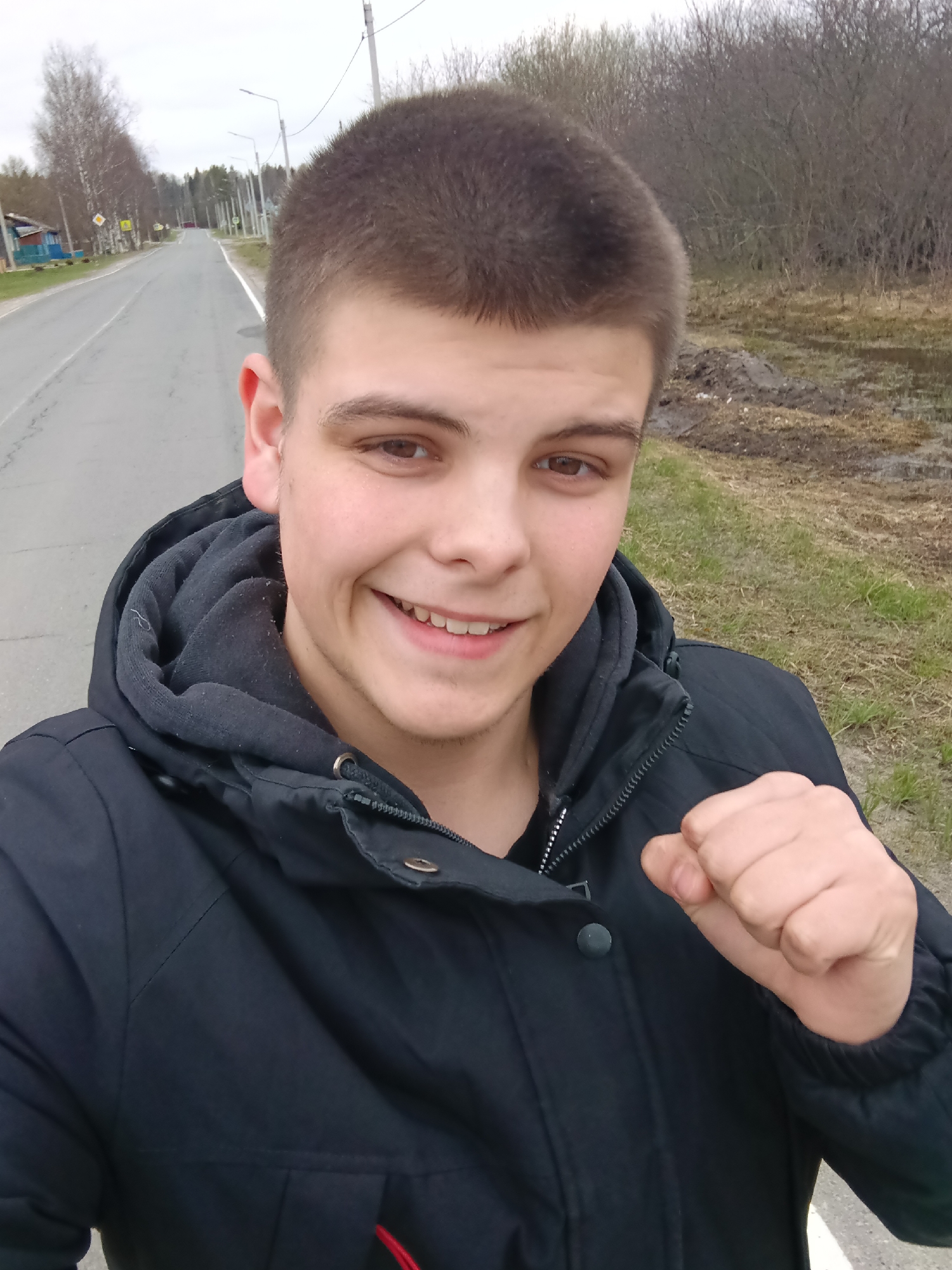 A young man is taking a selfie on a road in Красный Маяк, Russia. He is wearing a black hooded jacket and smiling at the camera, with his right hand clenched in a fist. A paved road runs along the left side of the frame, with houses and trees visible in the distance. To the right is a grassy area and trees without leaves, suggesting either late autumn or early spring. The sky is overcast.