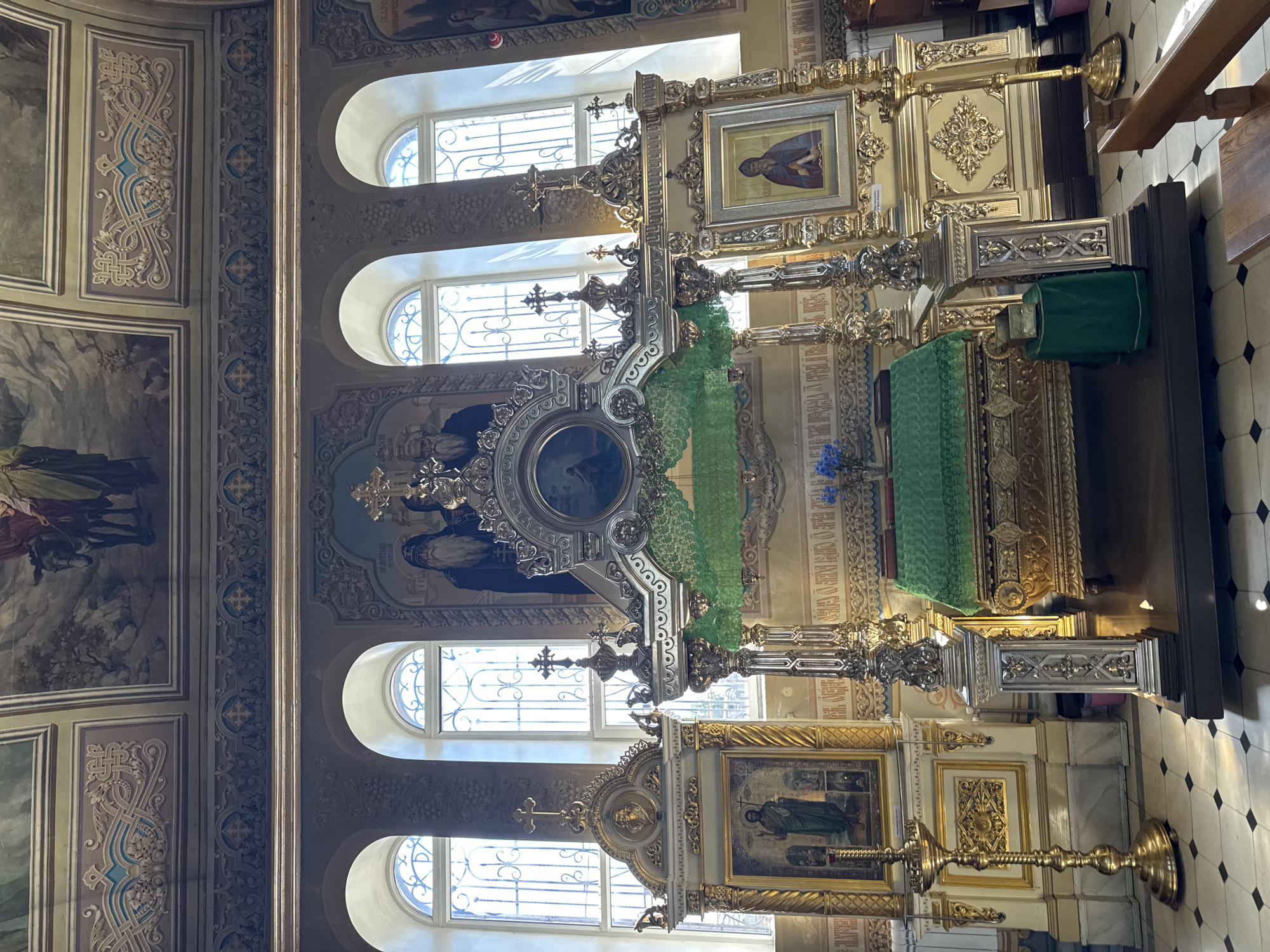 This is a medium shot, eye-level, interior photograph of an Orthodox church altar area. The scene is ornate and gilded. In the center is a large, elaborate altar structure with golden columns and decorative carvings. A green cloth is draped over a chest-like structure in the center, with a small bouquet of blue flowers on top. To the left and right of the central altar are smaller icon stands, also gilded, each displaying religious icons. Behind the altar are large arched windows with ornate grilles. Above the windows, the ceiling is decorated with paintings of religious scenes and intricate ornamental molding. The floor is tiled in a black and white diamond pattern. Sunlight streams through the windows, casting shadows on the floor. There are no people visible in the frame. The overall impression is one of solemnity and grandeur.