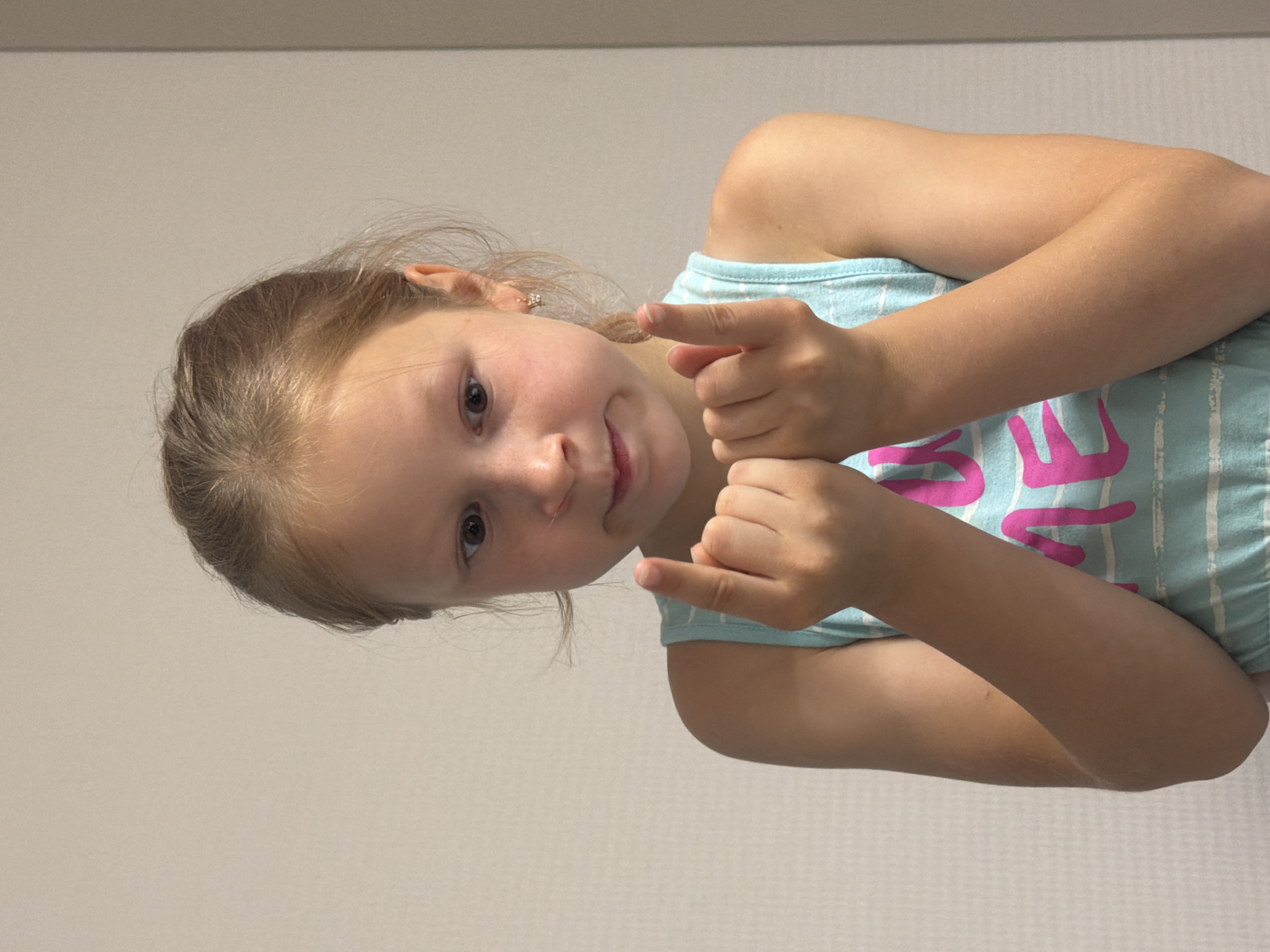 This is a media file of a young girl, likely a child, with blonde hair and fair skin. She is wearing a light blue tank top with white stripes and pink text that is partially obscured. Her facial expression is somewhat playful, with a slight smile and her eyes looking towards the viewer. She is holding up her index fingers, pointing them outwards in a gesture that could be interpreted as playful or directive. The background is a plain, light-colored wall, suggesting an indoor setting. The lighting appears to be soft and diffused, indicating it might be daytime or an interior with good lighting. There are no clear indicators of the weather or time of day. The context of Dubovaya, Russia, doesn't provide specific visual cues within the image itself.