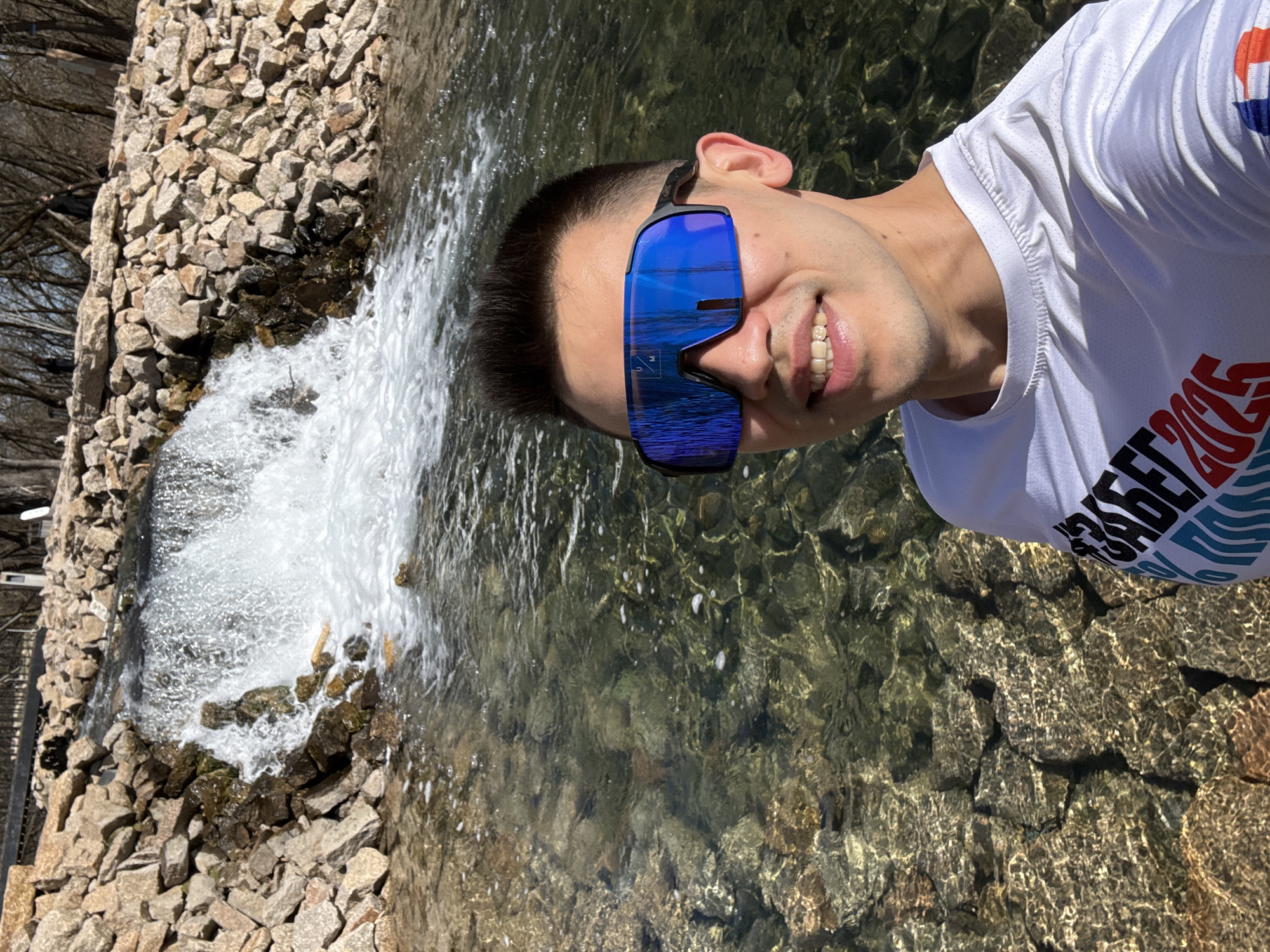 A man in a white shirt and blue sunglasses stands near a waterfall, possibly for a selfie or photo. The water cascades over light-colored rocks. The water reflects the sunlight and the rocks below. Trees and a building are visible in the background. Based on the context clues in the clothing print, it is likely this is a photo from Kazan, Russia.
