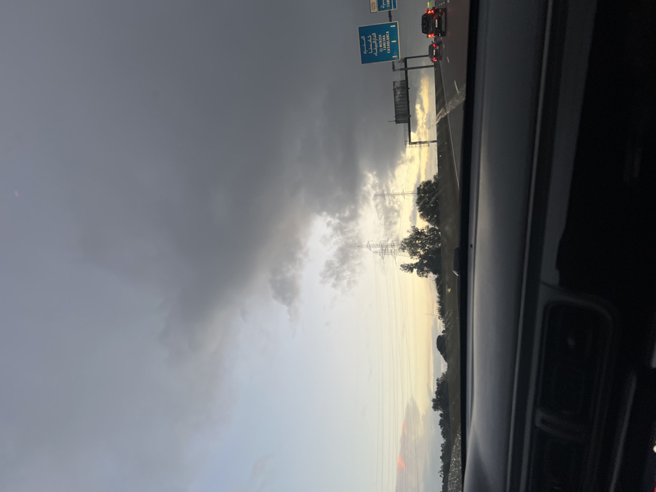 The photograph shows a cloudy sky from inside a vehicle. Dark storm clouds fill the left side of the frame. Power lines and a metal transmission tower appear in the distance on the right, with trees below. A road sign is visible above the car in front. The overall light suggests it may be either sunrise or sunset.