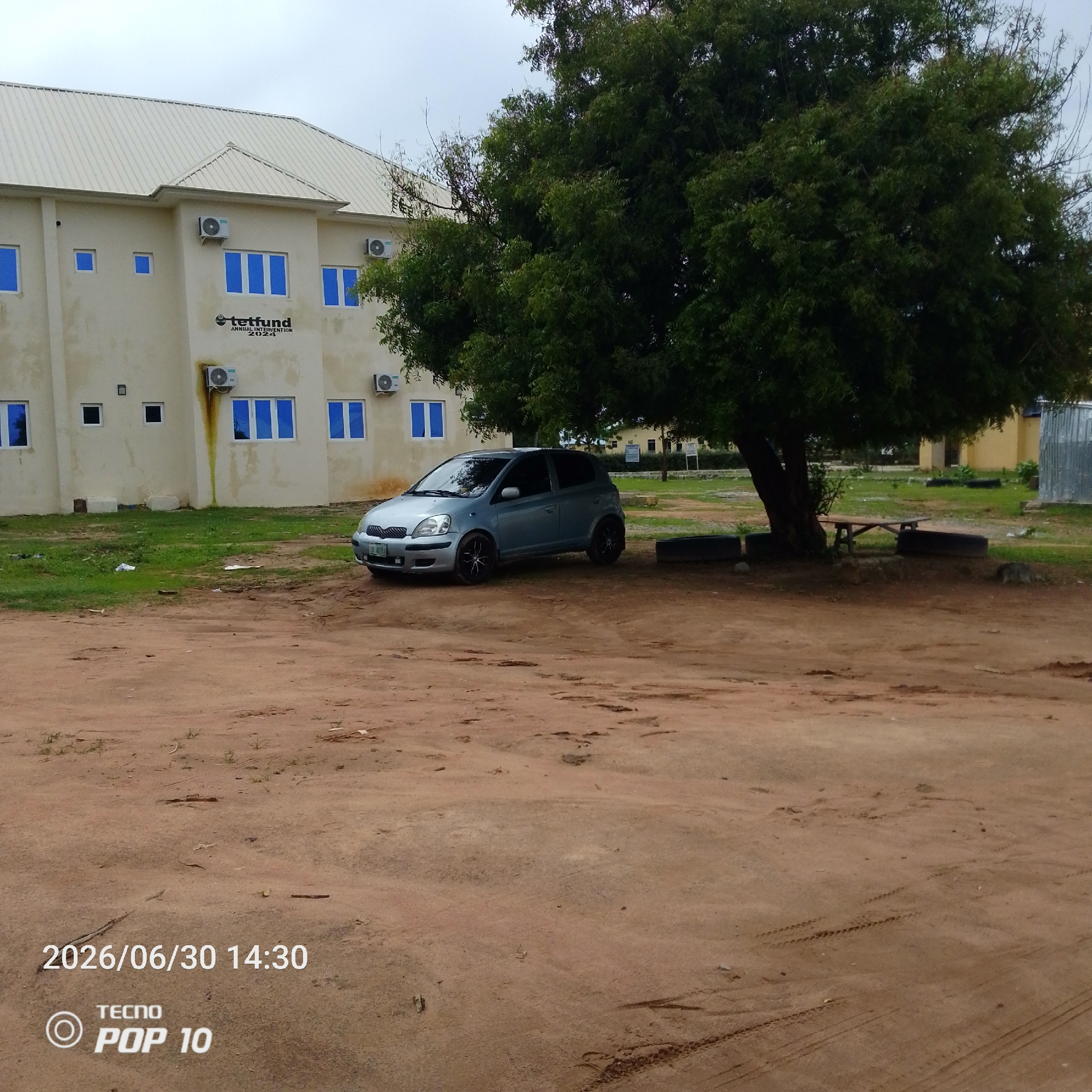 The image depicts an outdoor scene in Jalingo, Nigeria, under an overcast sky. In the background, a light-colored, multi-story building with a light-colored metal roof is visible. The building has several windows with blue-tinted glass and air conditioning units mounted on its exterior. A sign on the building reads "tetfund ANNUAL INTERVENTION 2024".
In the foreground, a silver hatchback car is parked on a dirt or gravel lot. The car has black rims with a white accent design. Beside the car and to the right, a large, leafy green tree provides shade. Further to the right, there's a wooden bench surrounded by what appear to be old tires, possibly repurposed as seating or decorative elements. The ground is predominantly dirt with patches of green grass and some scattered debris.
The overall atmosphere is calm and somewhat subdued, likely due to the cloudy weather. The timestamp "2026/06/30 14:30" is superimposed on the image, along with the "TECNO POP 10" logo. The scene appears to be a campus or an institutional setting given the building and its signage. No people are visible in the frame.