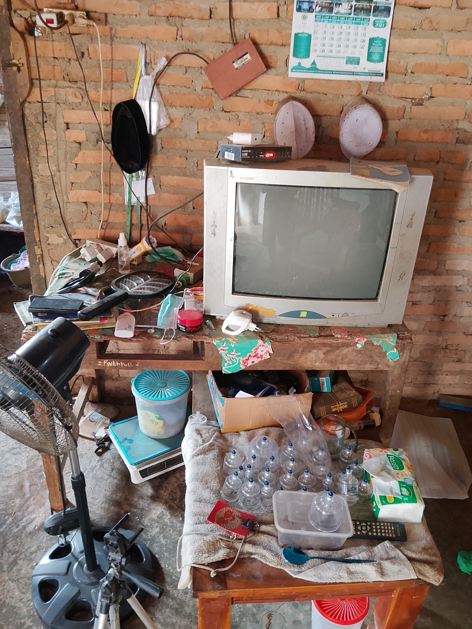 The image depicts an indoor scene, likely a cluttered living space or shop, in Negeri Agung, Indonesia. The environment is characterized by a rough brick wall in the background, a wooden table or shelf unit in the foreground, and a concrete floor.
The primary subject is an old, CRT television set placed on the shelf unit. Beside the television, on the wall, a calendar with religious imagery and text is displayed. Two white, oval-shaped caps, possibly prayer caps, are also visible on the wall near the calendar. Various electronic devices, including a fan and a remote control, are present. The shelf unit itself is laden with a diverse collection of objects, suggesting a state of disarray or a storage area. These include bottles of what appear to be medicinal or cosmetic products, glass cups, a box of tissues, a plastic container, a spoon, and other miscellaneous items. The presence of a black skullcap hanging on the wall further hints at a cultural or religious context.
The overall impression is one of a lived-in space with items accumulated over time, possibly indicating a home, a small business, or a storage area. The lighting appears to be natural and possibly supplemented by artificial sources, casting shadows that add to the depth of the scene.