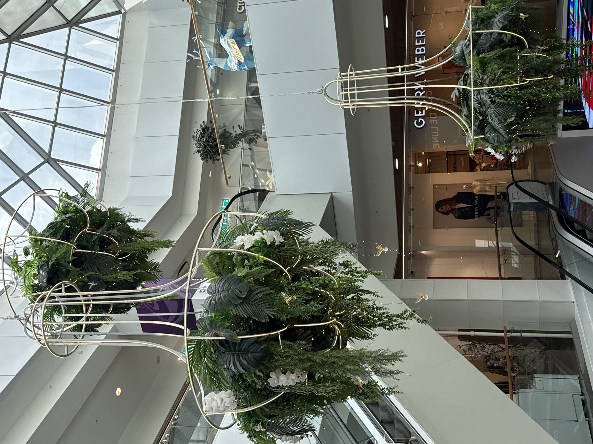 The image shows a wide-angle view of the interior of a shopping mall. Large, ornate chandeliers made of metal and filled with lush green plants and artificial flowers hang from the ceiling. The chandeliers are adorned with small, string lights. In the background, there are escalators, glass railings, store facades with signage including "GERRY WEBER," and a large window with a skylight. A decorative blue and white reindeer figure is visible on an upper level. The overall scene depicts a bright, modern, and well-decorated commercial space. The location is identified as the "Tachma" shopping mall in Izhevsk, Russia.