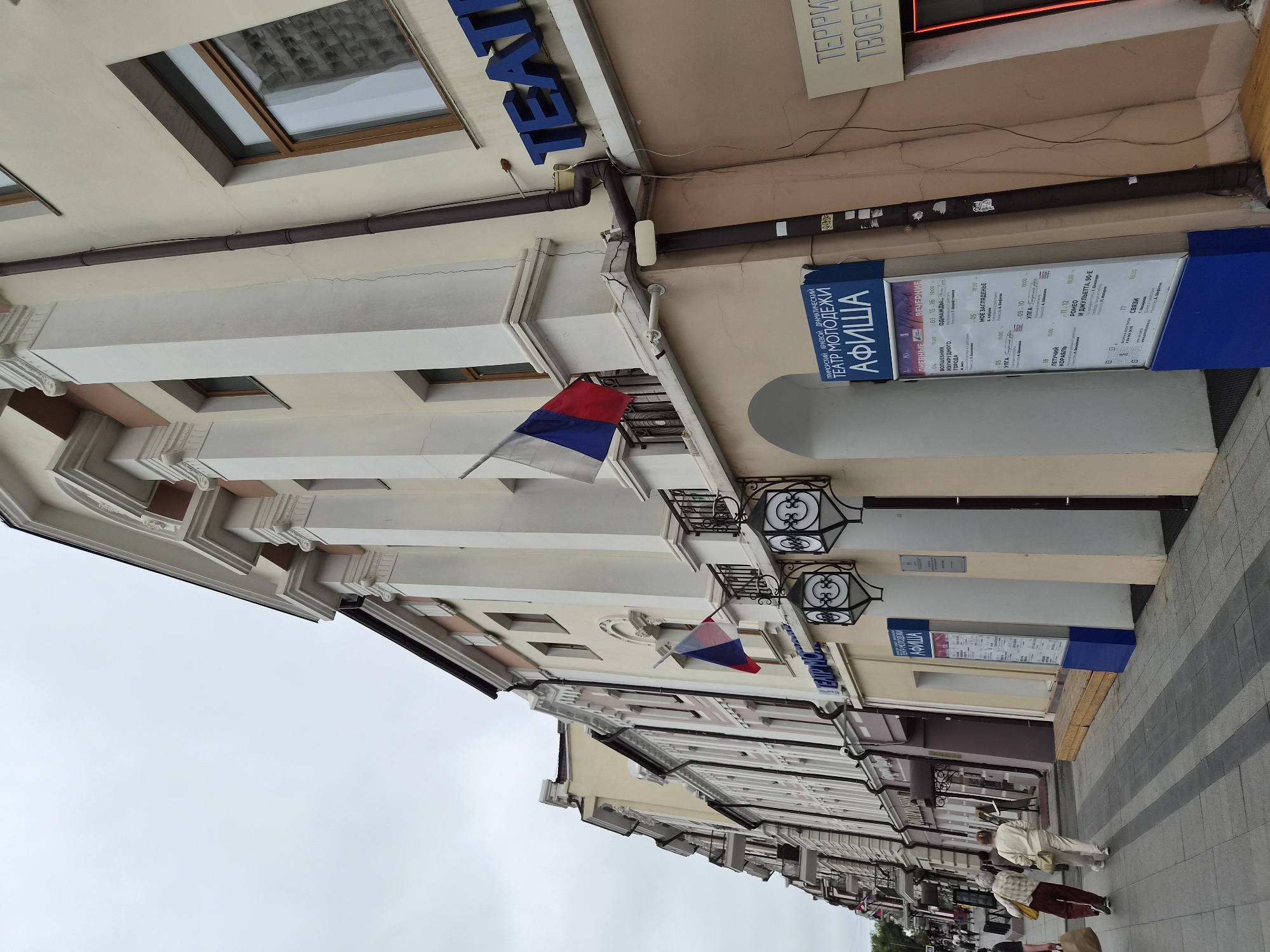 The image depicts a street scene in Vladivostok, Russia, specifically on Svetlanskaya Street. The focus is on a cream-colored building with classical architectural elements, including large columns and ornate moldings. Russian flags are displayed on the facade. To the left of the building, a row of other establishments with shops and balconies lines the street, indicating a commercial and residential area. In the foreground, two elderly women are walking on the sidewalk. A blue informational kiosk, labeled "ТЕАТР МОЛОДЕЖИ" (Youth Theater), displays a schedule of events, suggesting the building houses a cultural venue. The sky is overcast, and the overall atmosphere is one of a typical urban day.