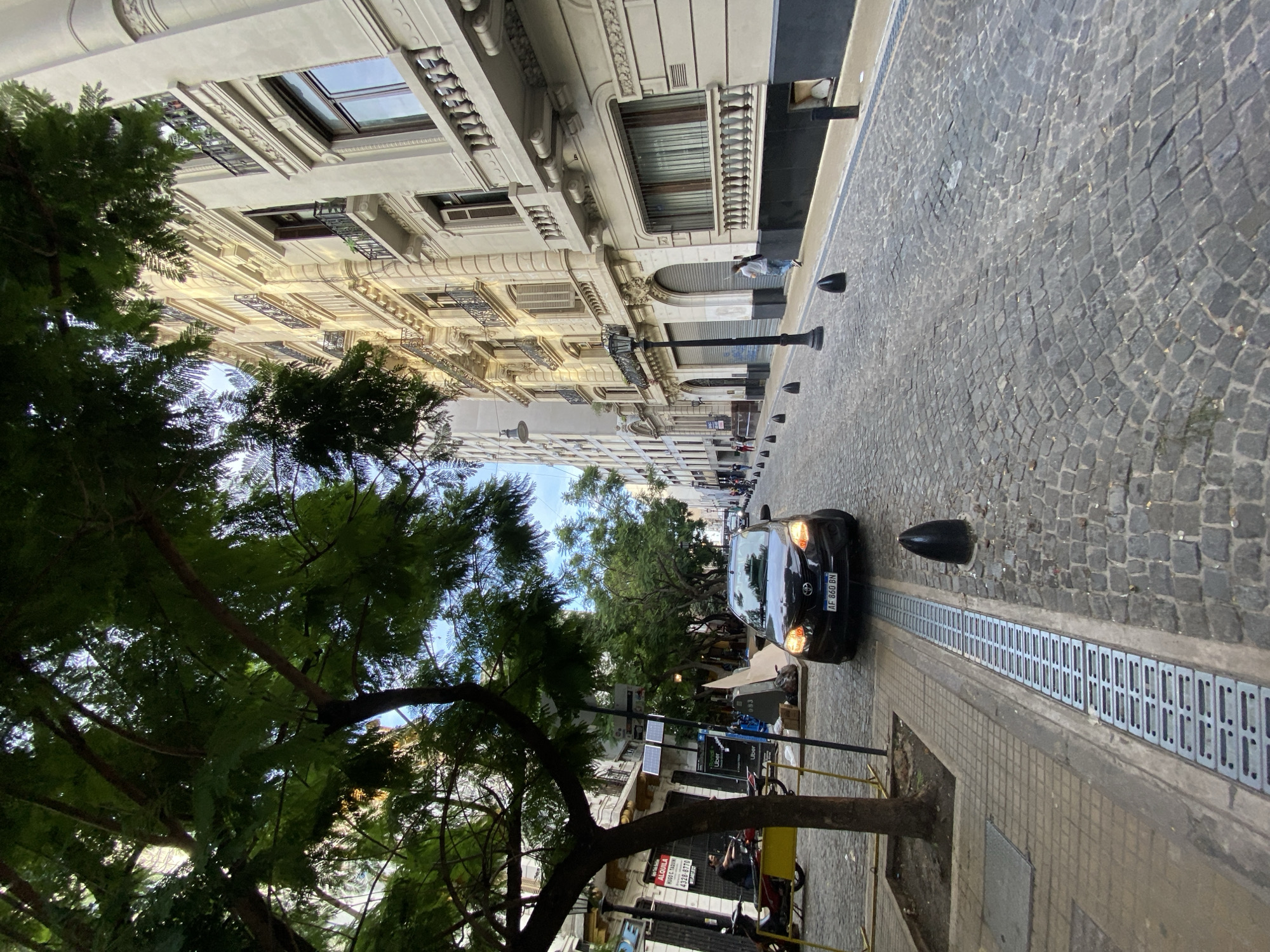 The image shows a street scene in Buenos Aires, Argentina. The camera is pointed down a street paved with cobblestones, lined with ornate, multi-story buildings on one side and a tree-lined sidewalk on the other. A black car, registered with the license plate "AF 860 BN", is driving down the street. A metal grate runs along the edge of the sidewalk, separating it from the street. There are several parked bicycles along the sidewalk. A person is walking on the other side of the street. Street lights are interspersed between the buildings. The sky is visible between the buildings and trees.