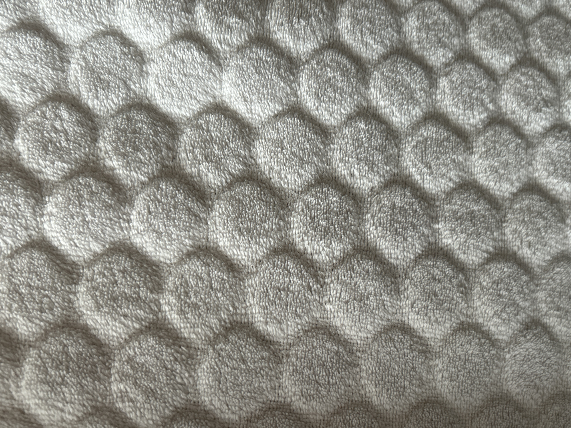 The image is a close-up, top-down view of a textured, light gray fabric with a hexagonal quilted pattern. The fabric appears soft and plush, with a fuzzy surface. The pattern creates raised hexagonal areas interspersed with recessed channels, giving the material a three-dimensional appearance. The lighting casts subtle shadows, emphasizing the texture and contours of the fabric. The image does not contain any information about its location in Moscow, Russia, or any people, actions, or interactions.