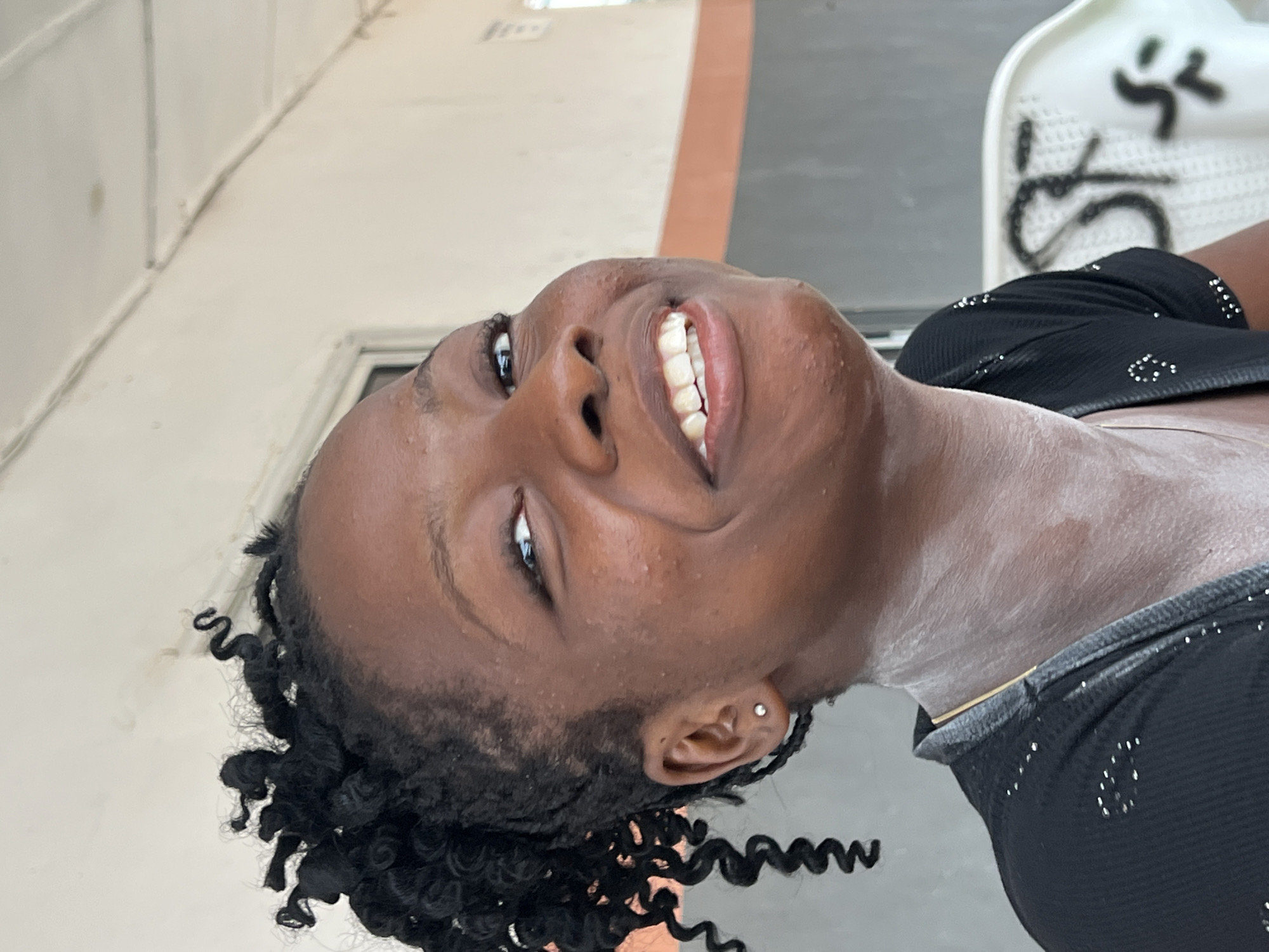 A young Black woman with dark skin, curly black hair styled in a bun, and a small stud earring in her left ear is smiling at the camera. She is wearing a black top with silver embellishments and has white powder on her neck. The background appears to be an outdoor setting with a light-colored wall and some indistinct structures. The image was captured in Nkpor, Nigeria.
