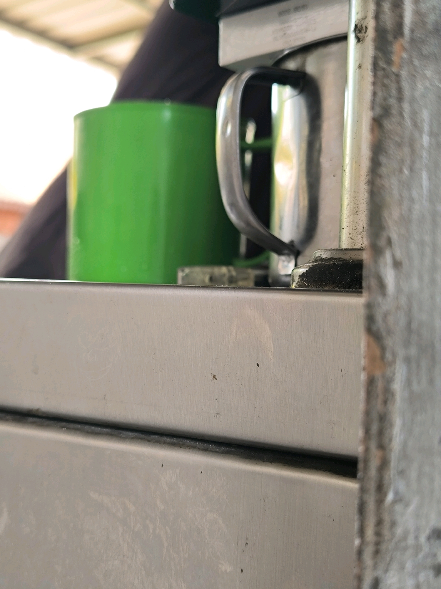 The image shows a close-up, low-angle view of a metallic structure, possibly a counter or appliance, in what appears to be an outdoor or semi-outdoor setting with a blurred background of a white roof and dark fabric. On the left, a bright green cylindrical object, likely a cup or container, is visible. To the right of the green object, a silver metallic handle is attached to a larger, reflective metallic surface. The metallic surfaces in the foreground appear slightly dusty or worn, with a darker seam running horizontally across the lower portion of the image. The lighting suggests it is daytime, with soft light illuminating the scene. No people are visible in the frame.